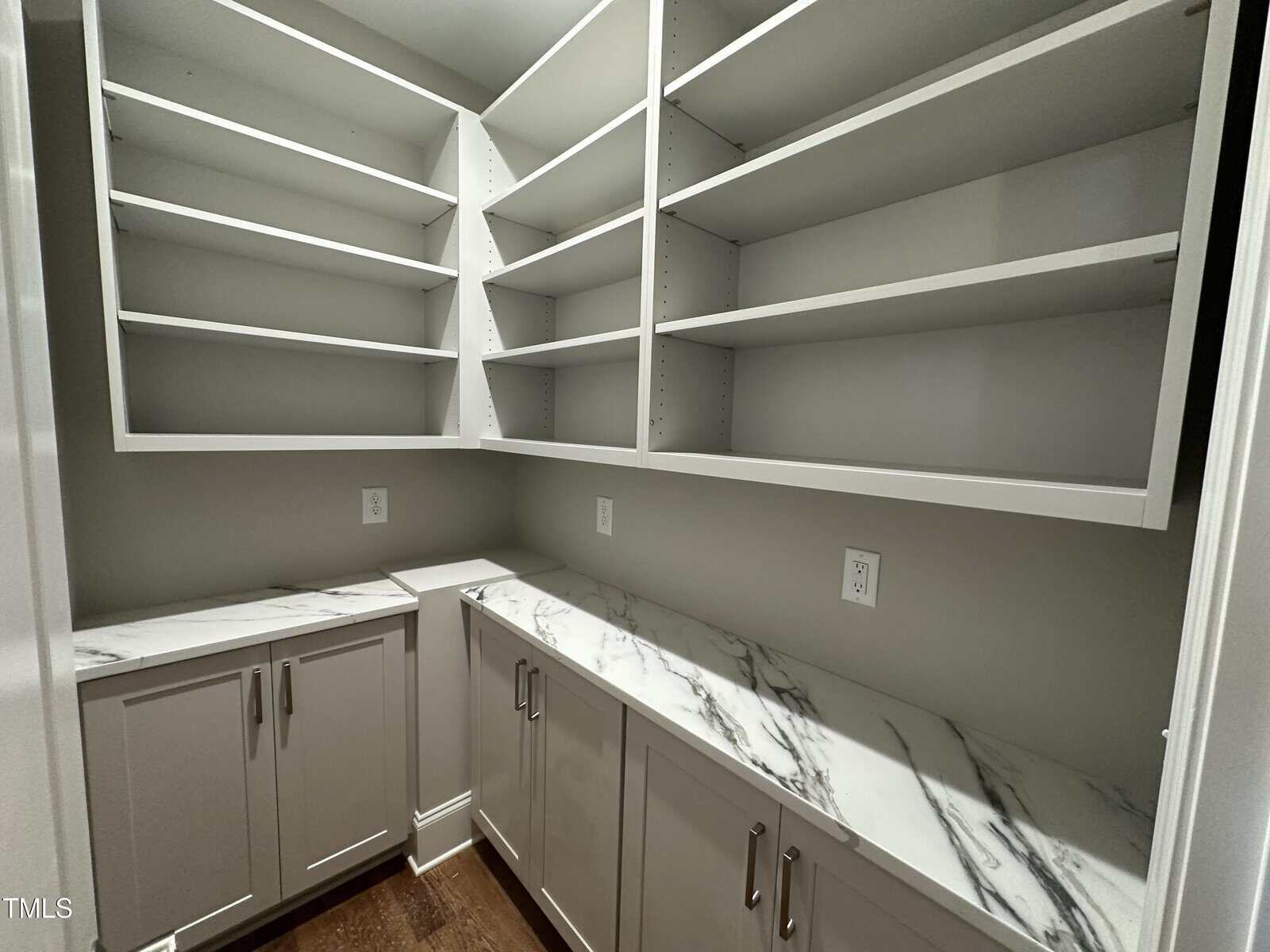 A walk-in pantry with white shelves on two walls, white lower cabinets with silver handles, and marble countertops. The walls are light gray, and the floor is dark wood. There are electrical outlets above the counters.