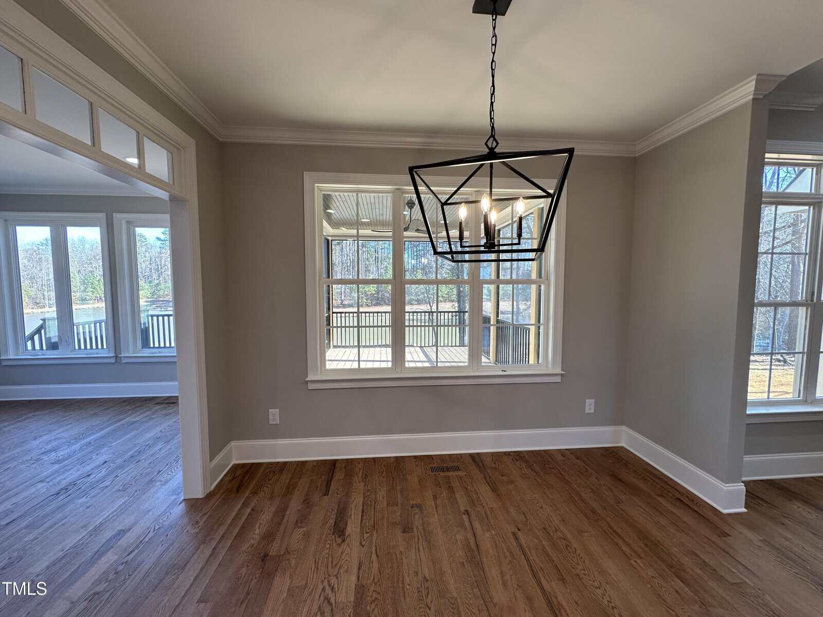 A dining area with hardwood floors, beige walls, and a modern black geometric chandelier. Large windows provide natural light, and an adjacent room with more windows is visible to the left.