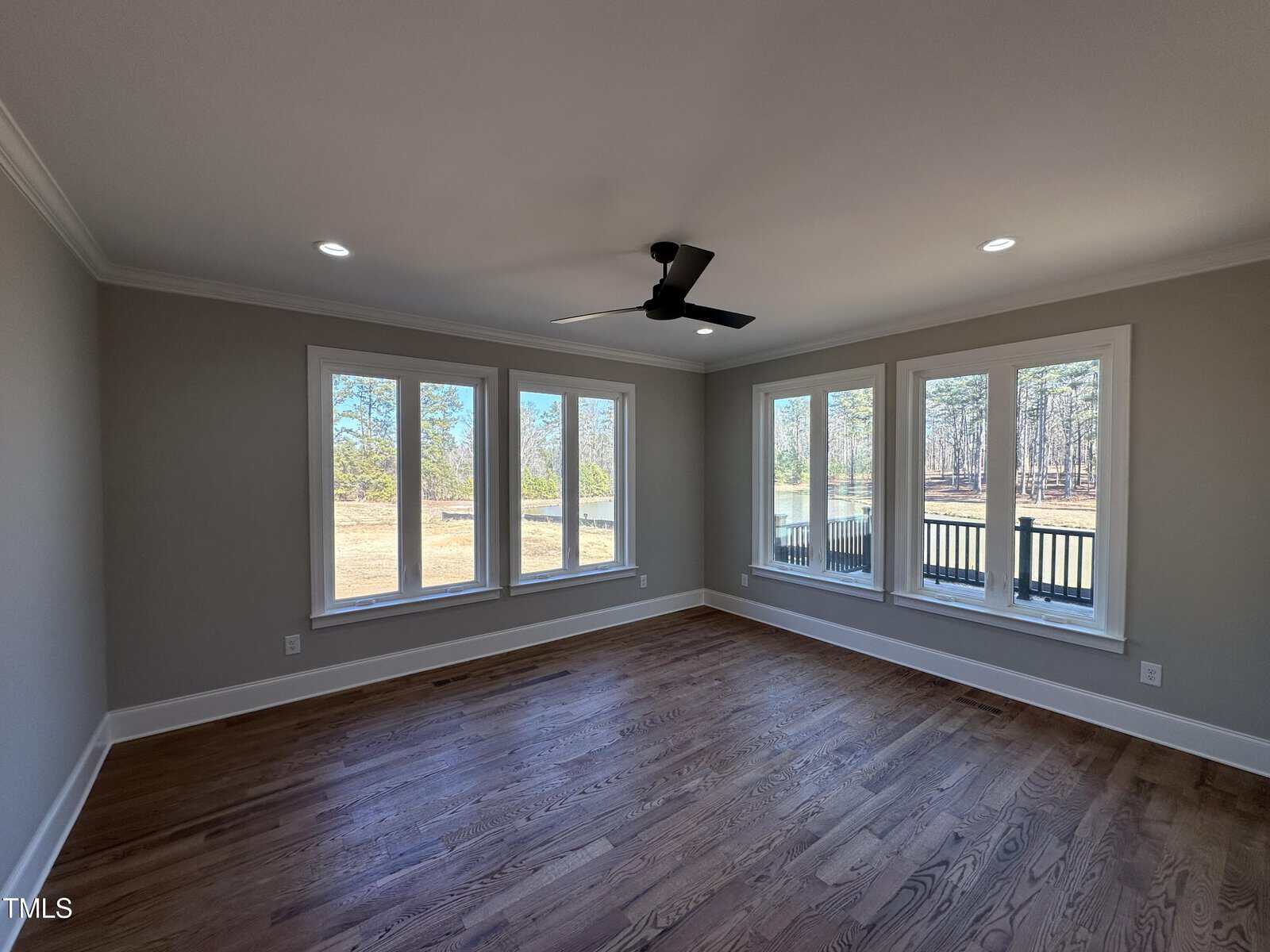 Empty room with hardwood floors, light gray walls, recessed ceiling lights, a black ceiling fan, and four large windows offering views of trees and a grassy yard. Natural light fills the space.