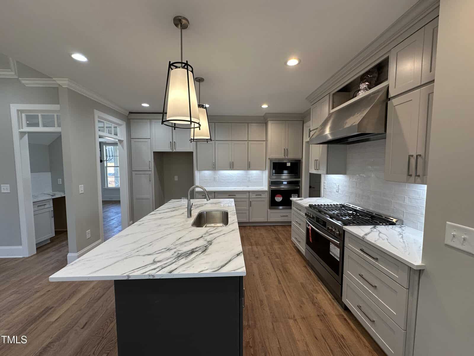 Modern kitchen with white cabinets, marble countertops, a large island with a sink, stainless steel appliances, gas range, built-in oven, wooden floor, and pendant lights hanging from the ceiling.