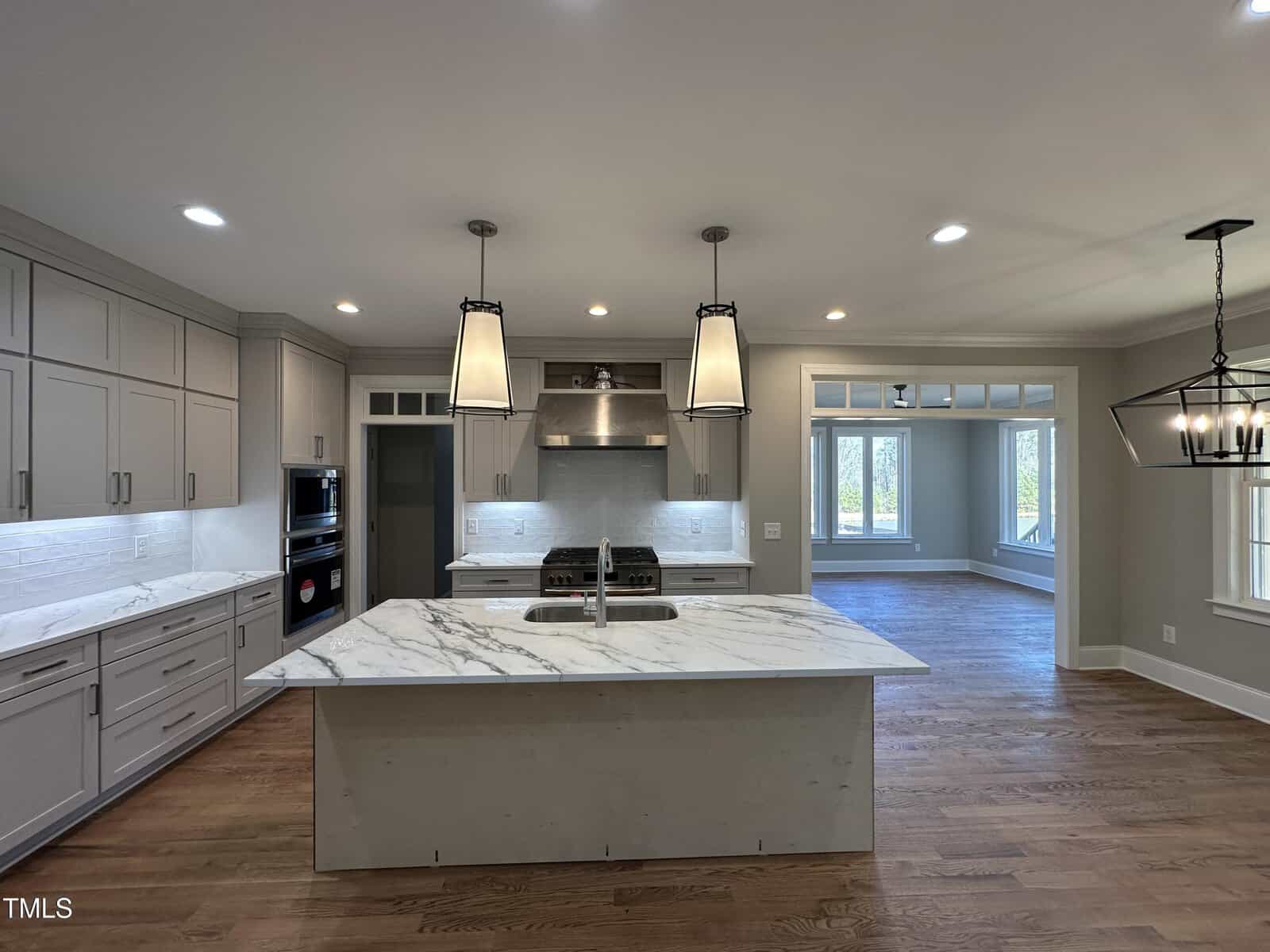 Modern kitchen with light gray cabinets, marble island, stainless steel appliances, and two pendant lights. The space opens to a living area with large windows and hardwood floors throughout.