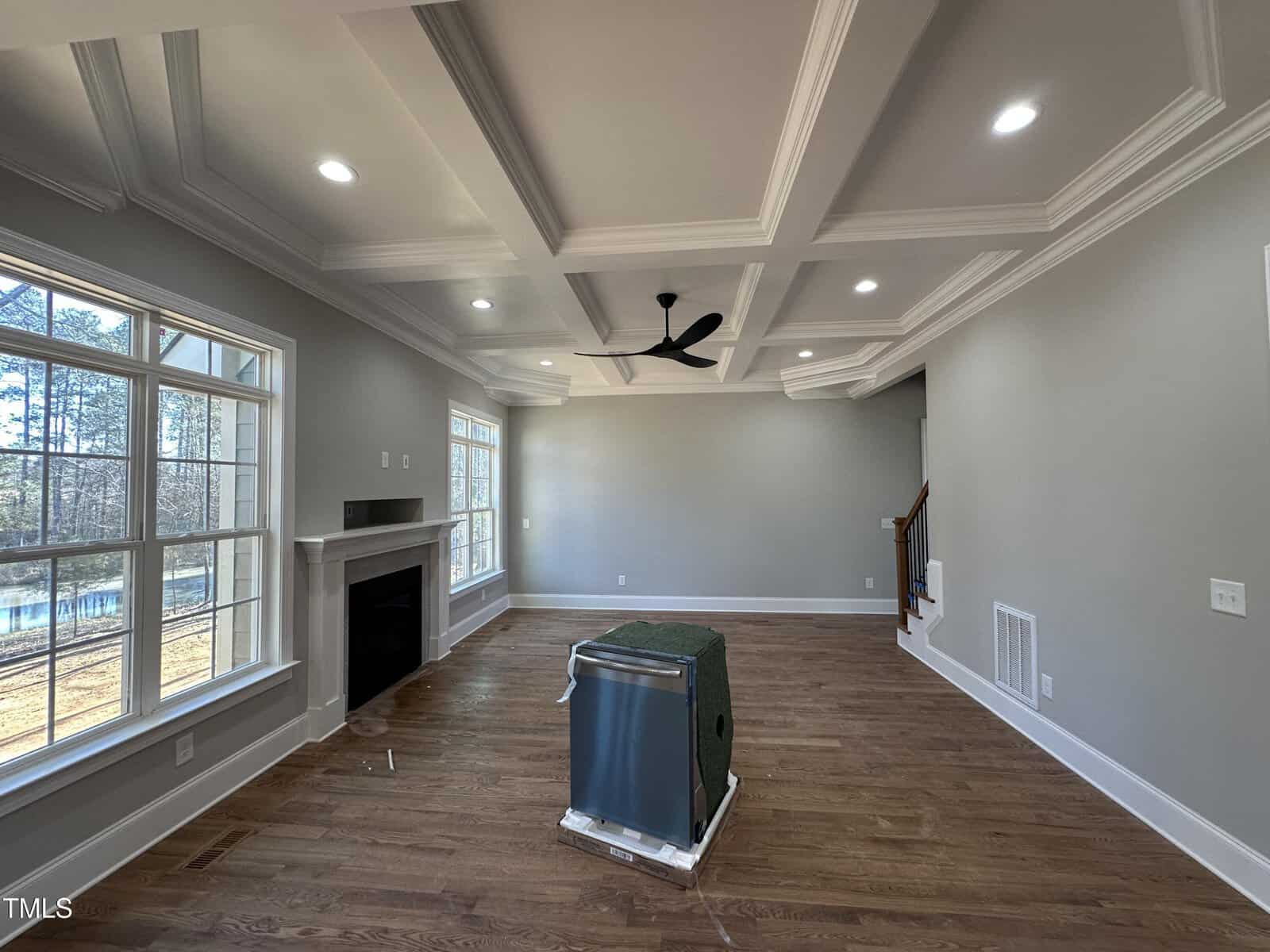 A spacious, unfurnished living room with large windows, a tray ceiling with recessed lighting, a modern ceiling fan, a fireplace, and hardwood floors. An uninstalled appliance sits in the center of the room.