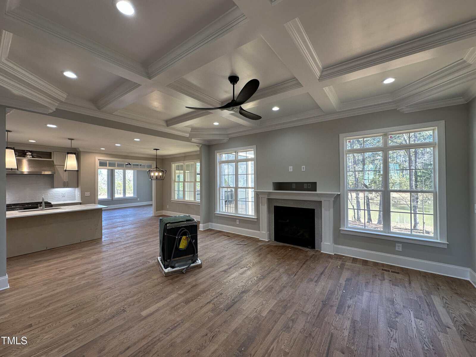 A spacious, open-concept living area with hardwood floors, large windows, a ceiling fan, recessed lighting, a fireplace, and an unfinished kitchen; a large portable fan sits in the middle of the room.