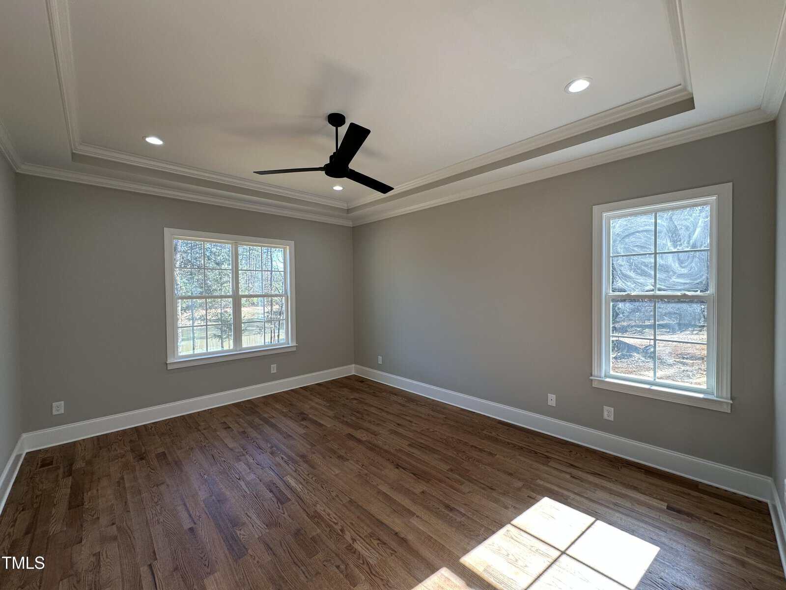 Empty room with light gray walls, wood floors, two windows, a black ceiling fan, and recessed lighting. Sunlight streams through the windows onto the floor.