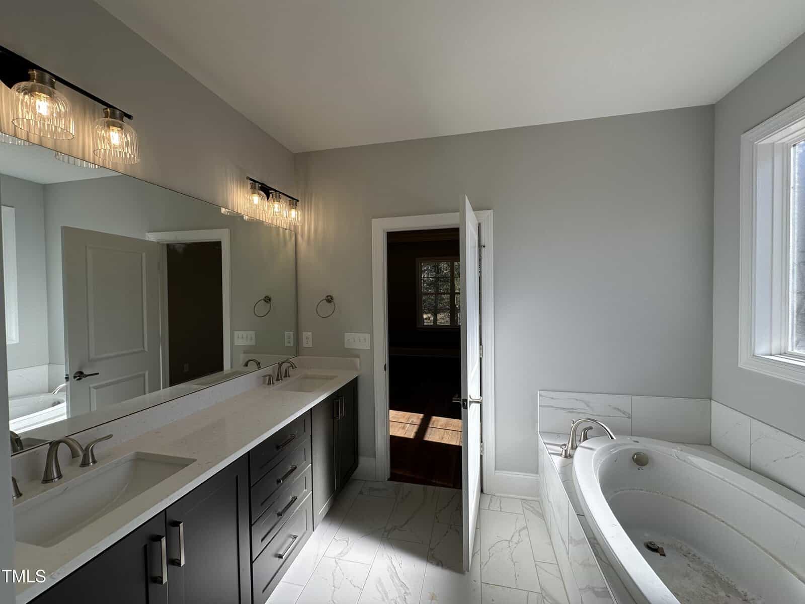 Modern bathroom with double sinks, dark cabinets, marble floors, a large bathtub, and two mirrors with contemporary lights above. Sunlight streams through a window and an open door reveals another room with dark floors.