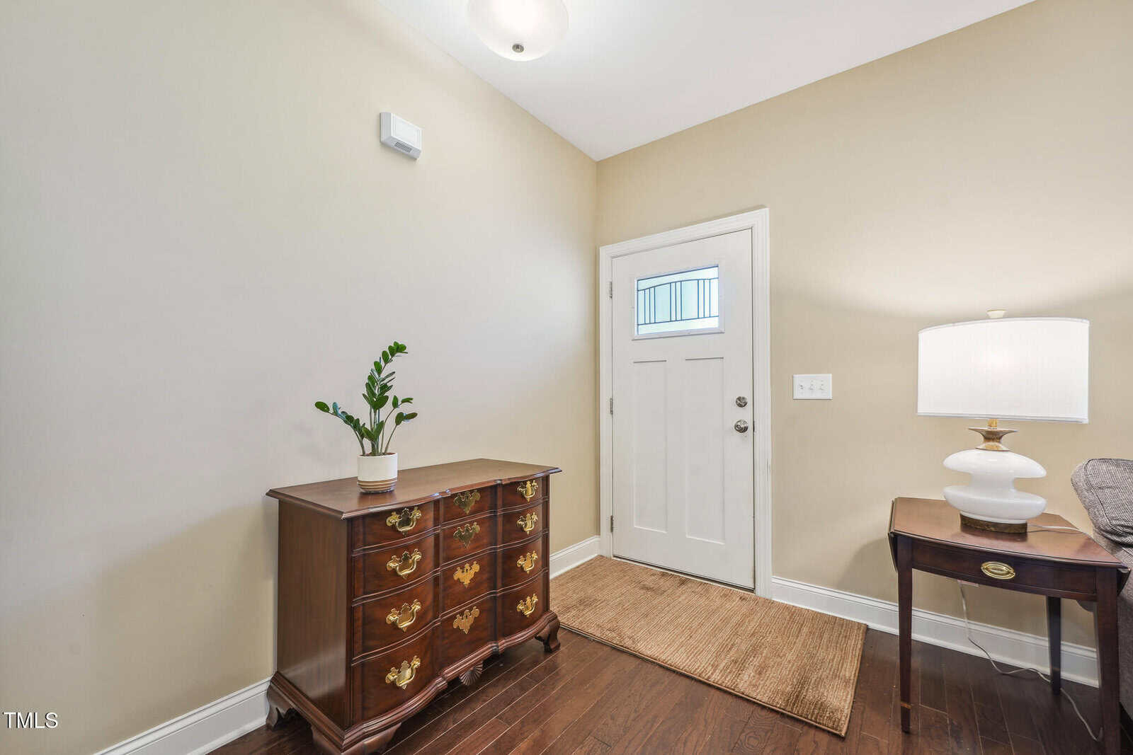 A small entryway features a white door with a glass panel, a wooden side table with a lamp and a beige sofa partially visible. A wooden chest with a potted plant sits against the beige wall on a dark wood floor with a rug.