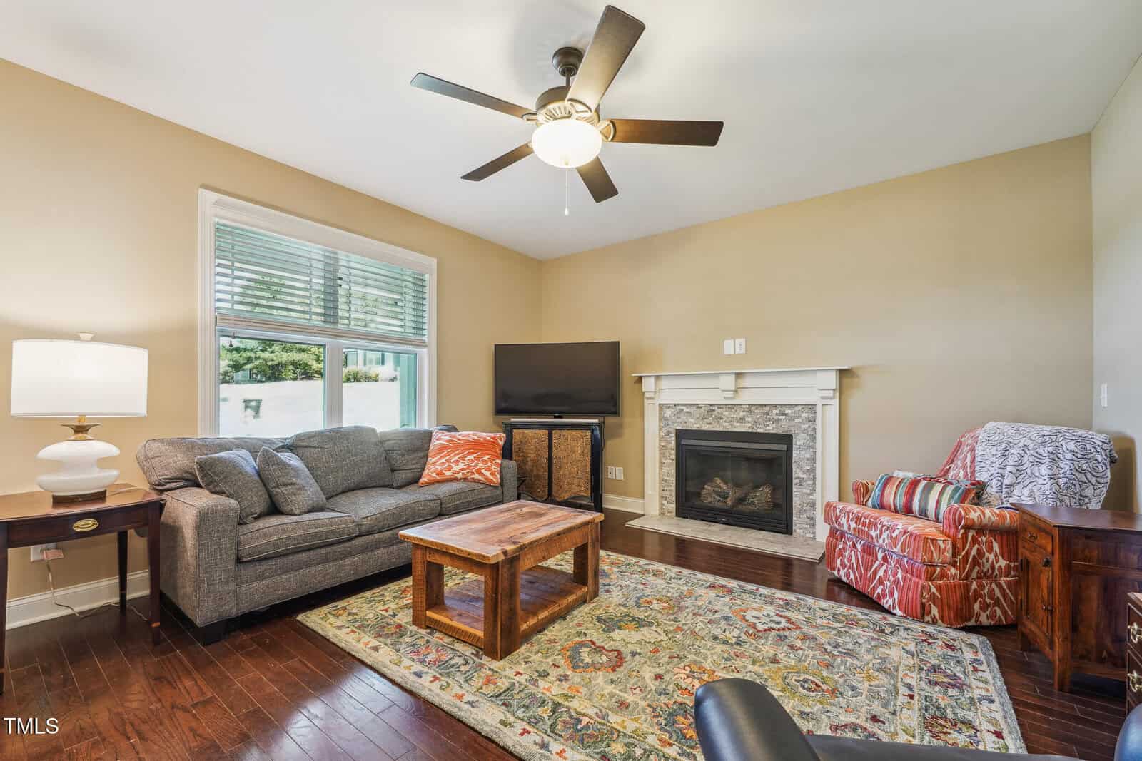 A cozy living room featuring a gray sofa, wooden coffee table, and patterned armchair. A fireplace with a mantel and a TV are set against a beige wall. A ceiling fan hangs above, and a colorful rug accents the hardwood floor.