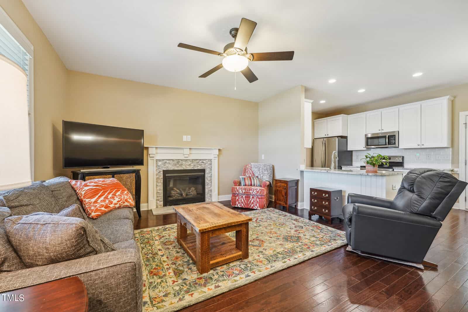 A cozy living room features a gray sofa, patterned armchair, and black recliner around a wooden coffee table. A fireplace is on one wall, with a TV nearby. The room opens to a kitchen area with modern appliances and a white countertop.