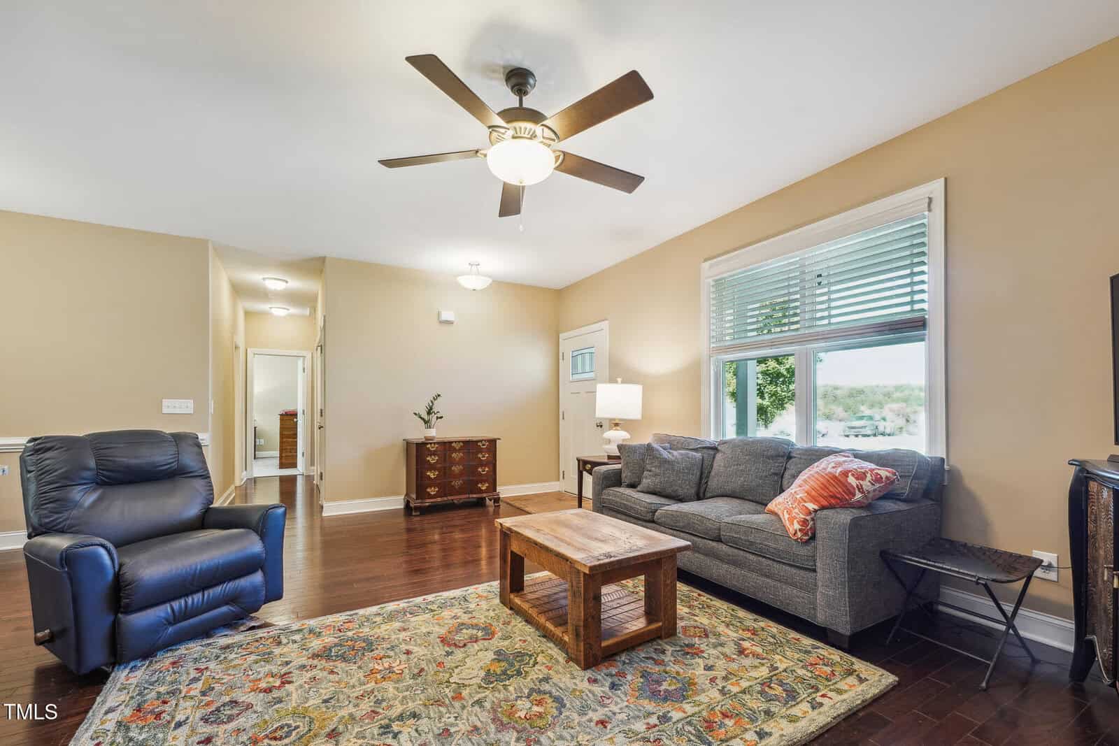 A cozy living room with a ceiling fan, dark wooden floor, a blue recliner, a gray sofa with orange cushions, a wooden coffee table, and a patterned rug. A window with blinds lets in natural light, and a wooden dresser is visible in the hallway.