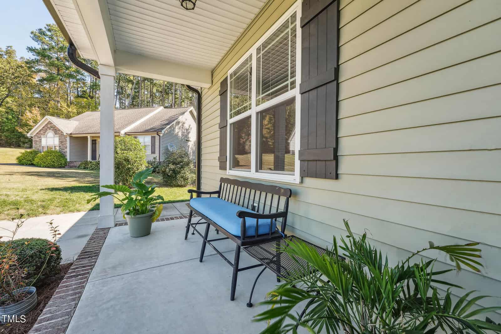 A porch with light green siding, featuring a black bench with blue cushions. Potted plants are arranged on the concrete floor. A house with similar siding is visible in the background under a clear sky.
