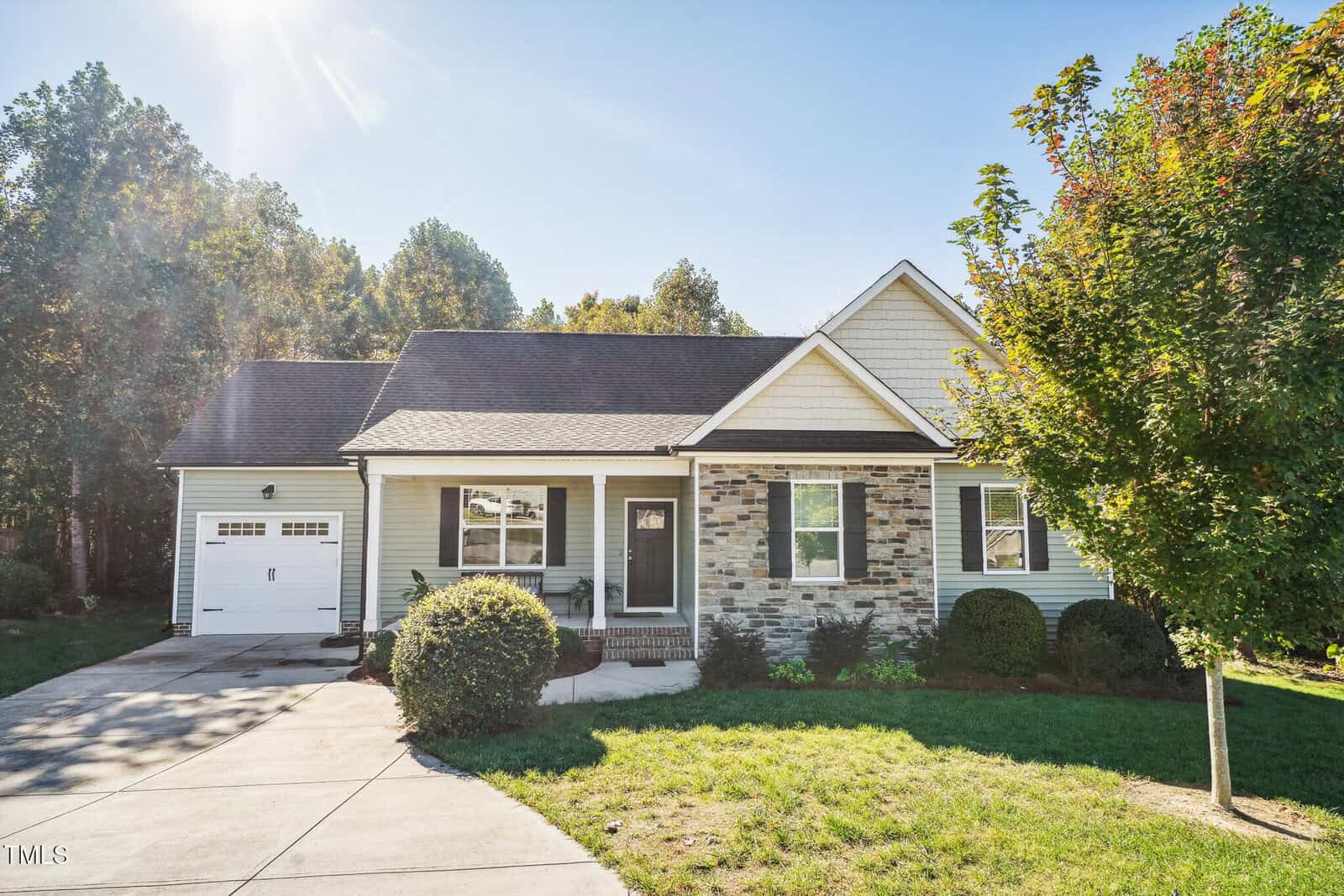 A single-story house with stone and siding exterior, featuring an attached garage and a small front porch. A well-maintained lawn and landscaped bushes are in the front. Sunny day with clear skies and trees in the background.