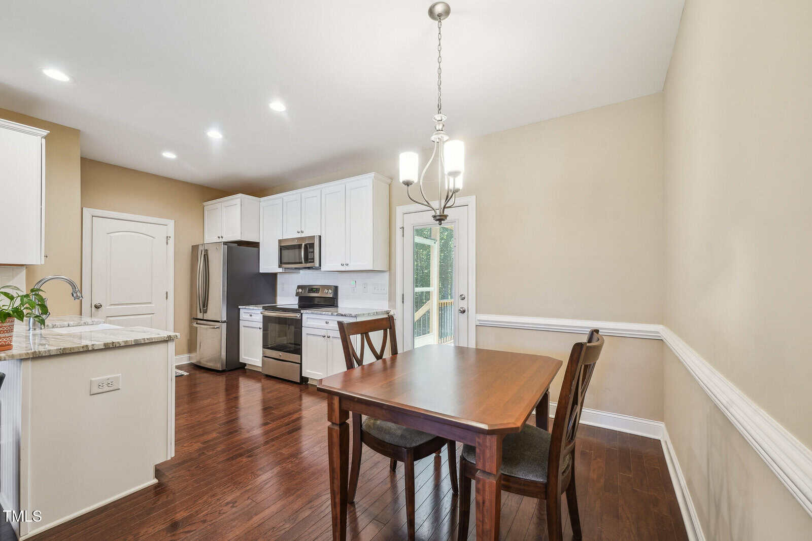 A kitchen with white cabinets, stainless steel appliances, and a wooden dining table with two chairs. A modern chandelier hangs above the table. The room has hardwood floors and a door leading to a backyard.