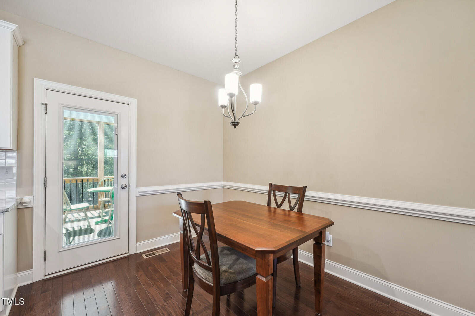 A cozy dining area with a wooden table and two chairs on a polished hardwood floor. A modern chandelier hangs above. A glass door opens to a view of a sunlit deck with trees visible through the windows. Walls are painted in a light beige.