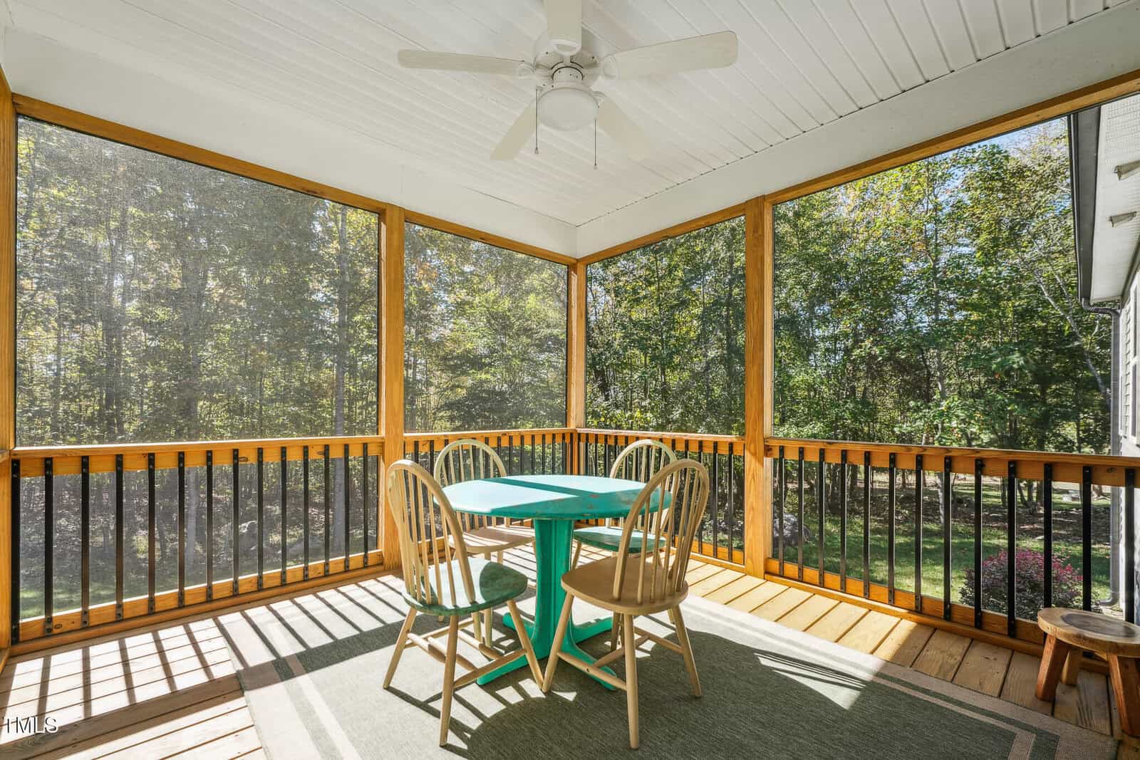Screened porch with a wooden floor, featuring a turquoise table and four wooden chairs. A ceiling fan is mounted above. Surrounded by trees, the porch offers a view of a lush, green landscape.