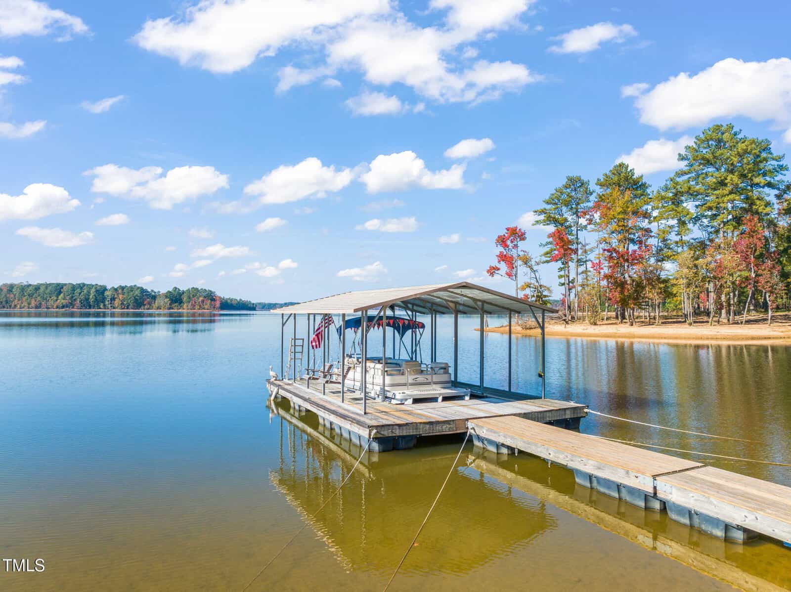 A covered dock with boats tied underneath sits on a calm lake, surrounded by trees with some autumn foliage under a blue sky with scattered clouds. A small walkway connects the dock to the shore.
