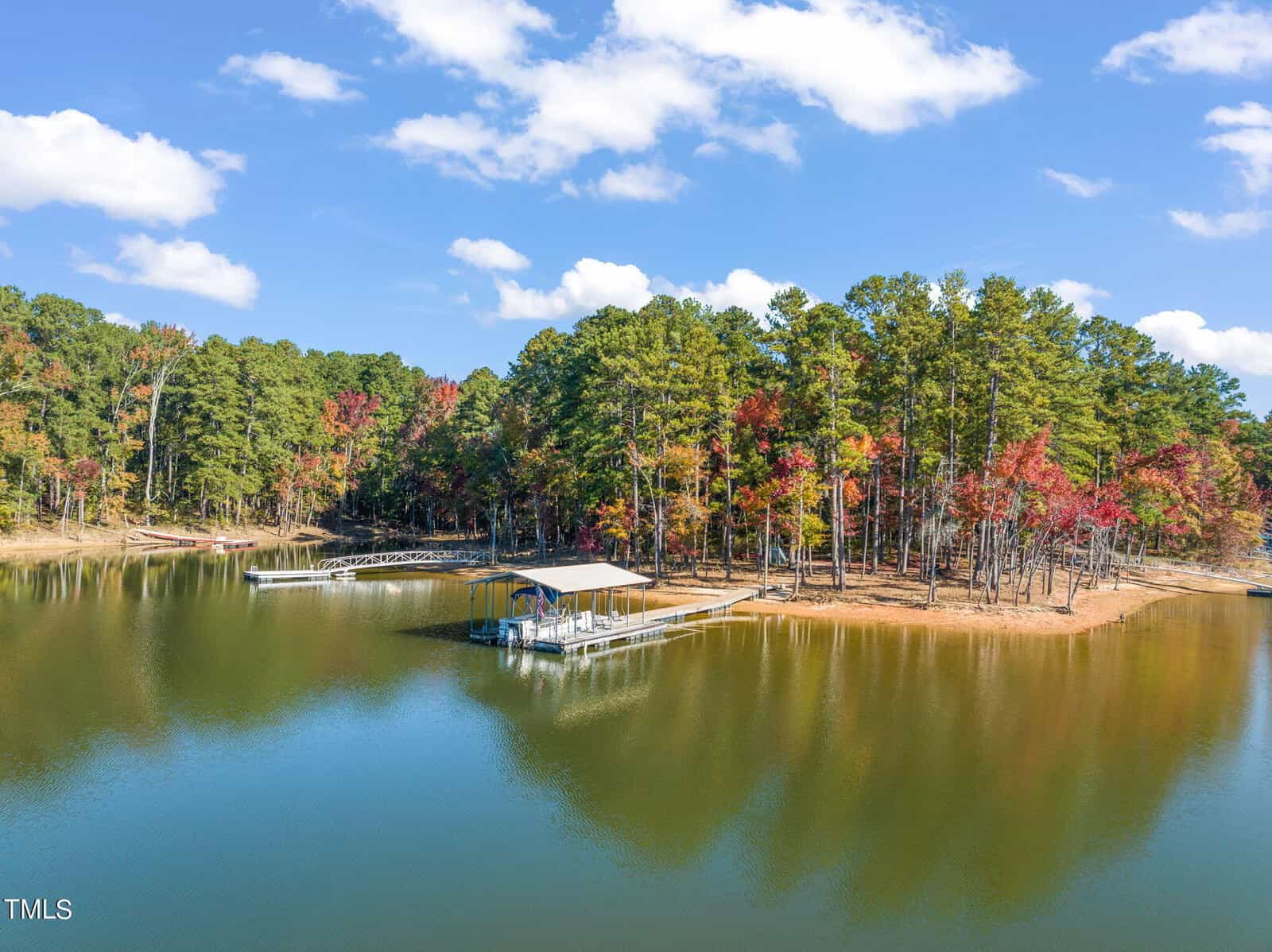 A peaceful lake with clear water reflects the sky and surrounding trees, some of which have autumn foliage. A covered dock extends into the water from a wooded shoreline under a blue sky with scattered clouds.