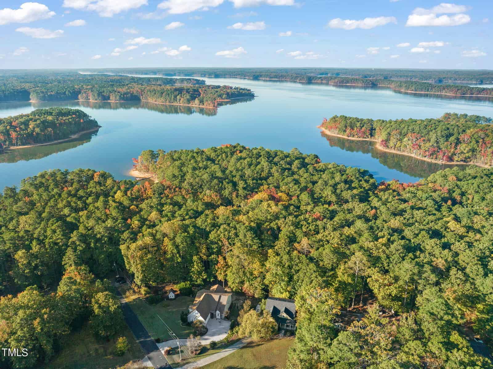 Aerial view of two houses surrounded by dense forest near a large, calm lake with small tree-covered islands under a blue sky with scattered clouds.
