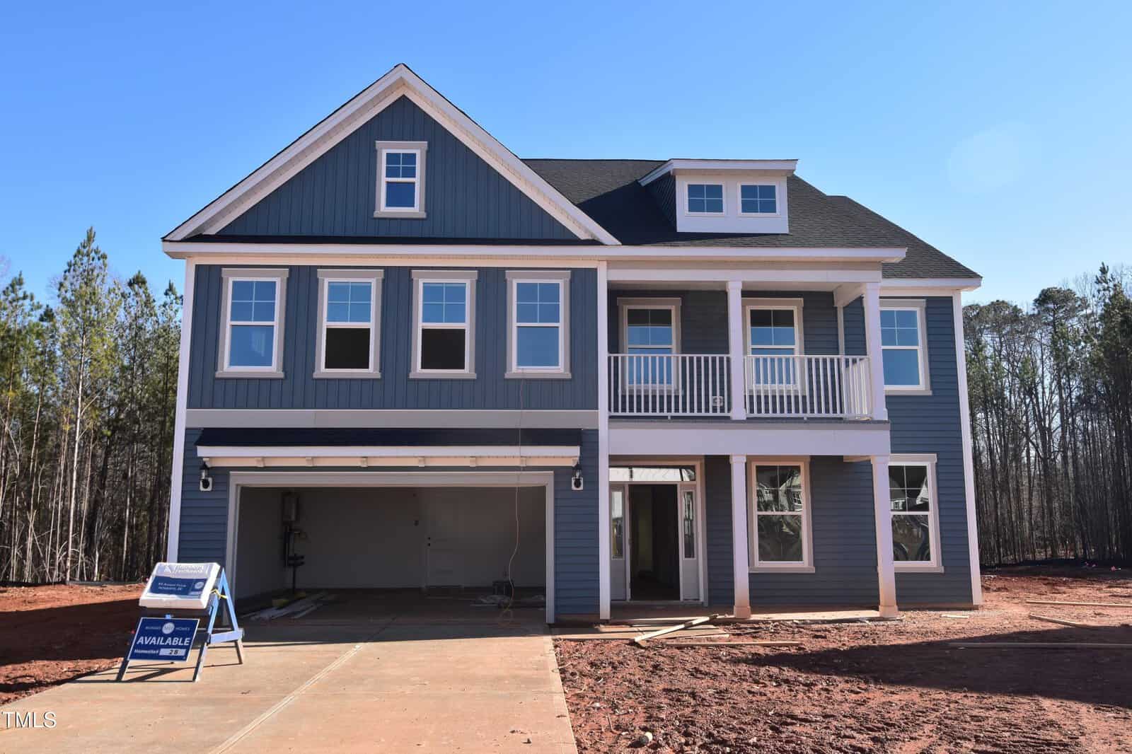 A newly constructed two-story blue house with white trim, double garage, multiple windows, and a second-floor balcony. A real estate sign indicating availability stands at the edge of the driveway.