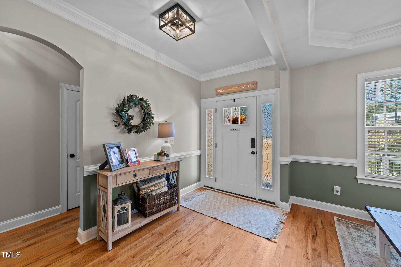 A bright entryway with wood floors, a white front door with glass panels, a side table decorated with photos and a lamp, a wall wreath, and sunlight streaming through windows with white blinds.