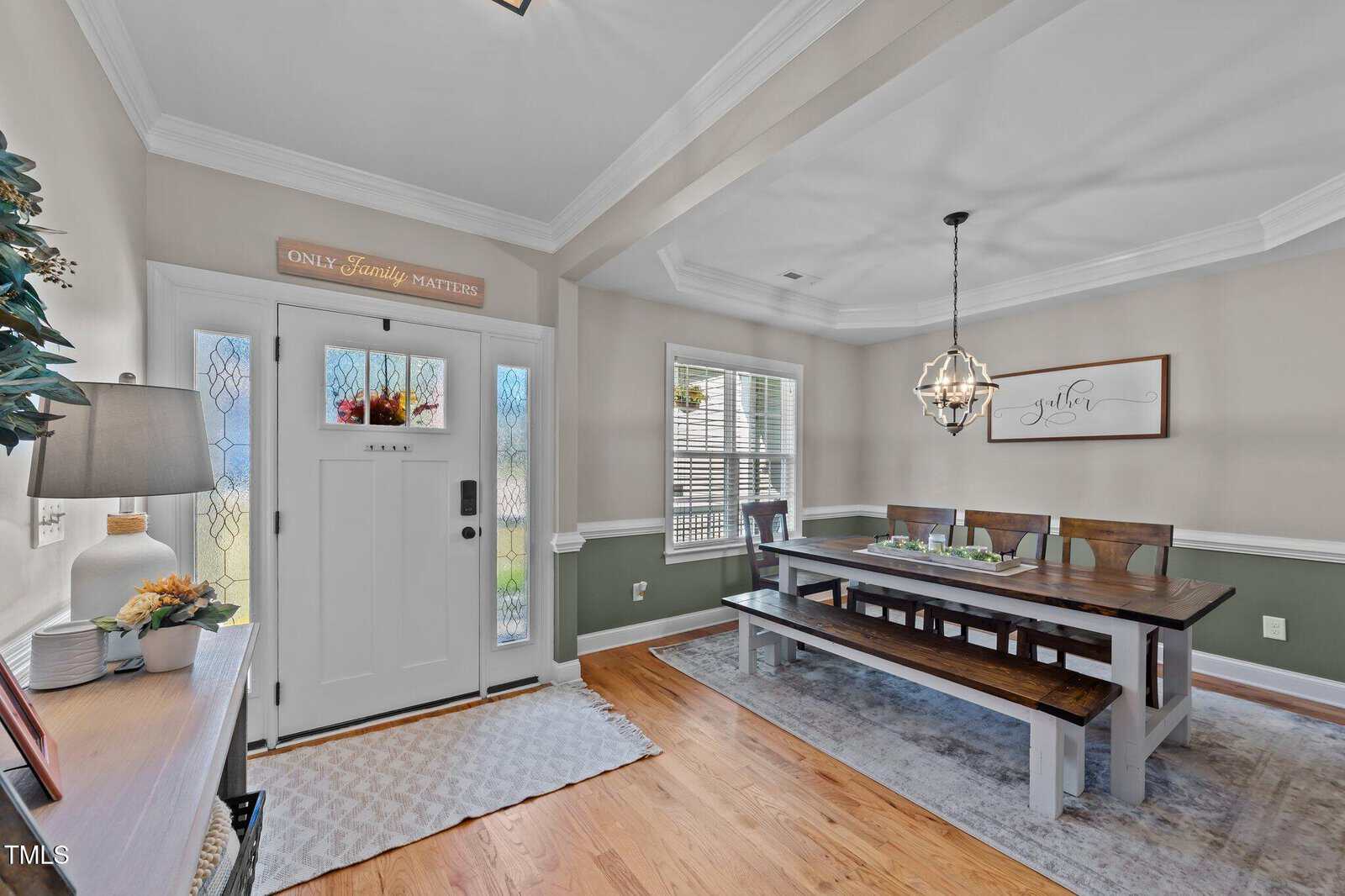 A bright dining room features a wooden table with benches and chairs, a chandelier, a sideboard with decor, hardwood floors, and a white front door with glass panels and a sign reading "Only Family Matters.
