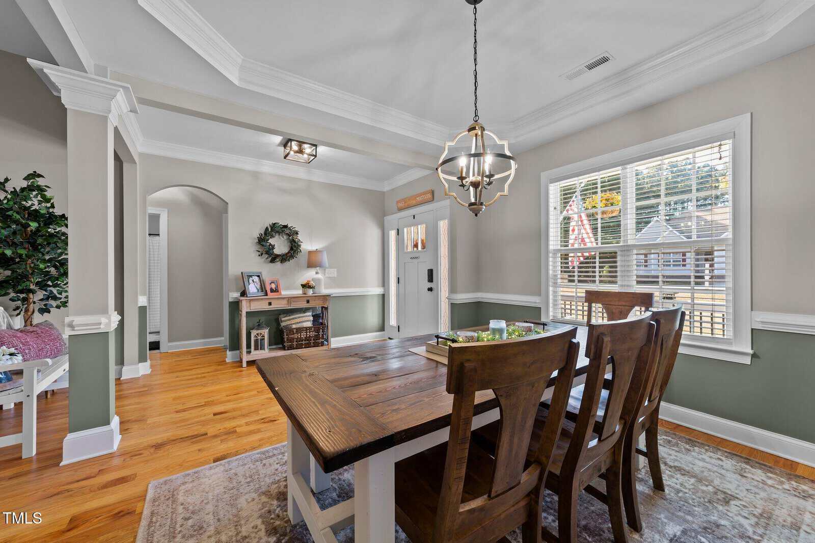 A bright dining room with a wooden table and four chairs, a large window with blinds, hardwood floors, light gray walls with green wainscoting, and a modern chandelier. A console table and decorative accents are visible.