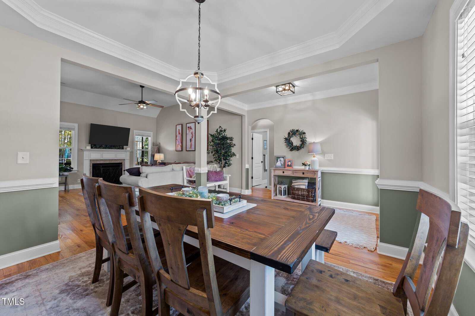 A bright dining room with a wooden table and six chairs, an overhead chandelier, hardwood floors, and light green lower walls. The room opens to a living area with a sofa, fireplace, TV, and decor elements.