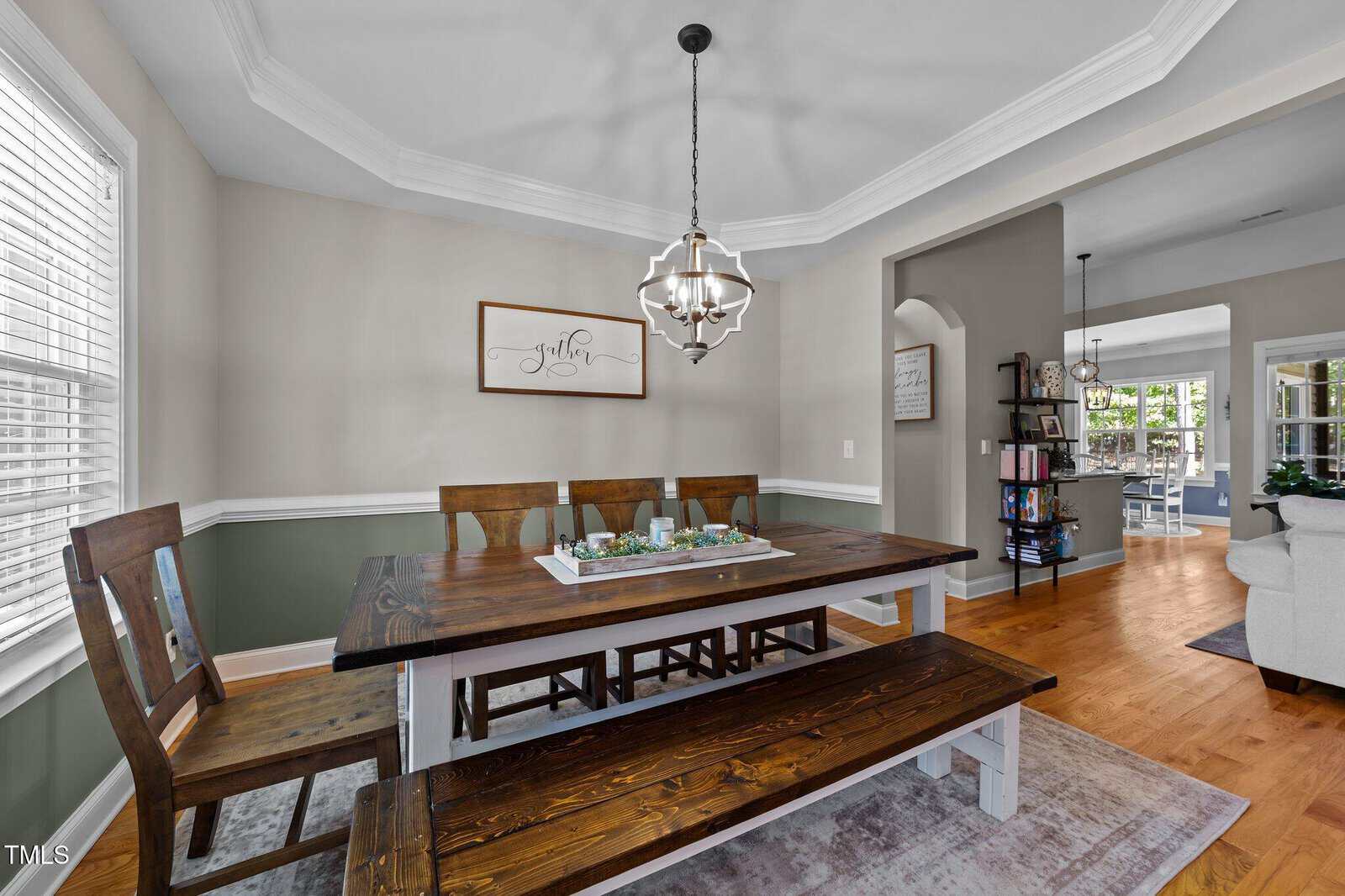A dining room with a wooden table, matching chairs, and a bench. A tray with decor sits on the table. The walls are two-toned with white trim, and a modern chandelier hangs above. An open doorway leads to a living area.