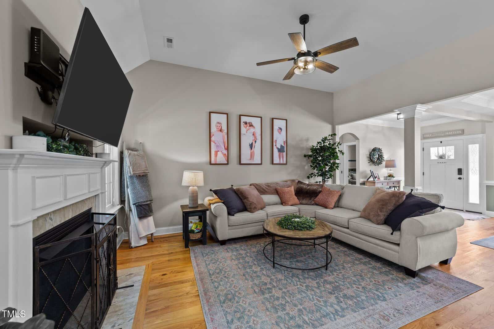 A cozy living room with a light sectional sofa, pink and beige pillows, a round coffee table, artwork on the wall, a fireplace, TV, ceiling fan, and a view of the front door entryway with natural light.
