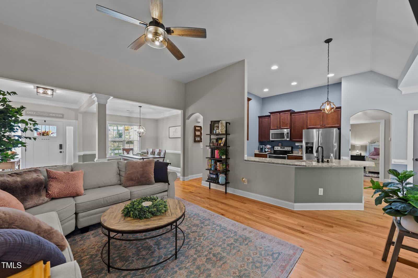 A cozy, open-concept living room and kitchen with hardwood floors, neutral walls, ceiling fan, sectional sofa, coffee table, bookshelves, and modern stainless steel appliances. Natural light brightens the space.