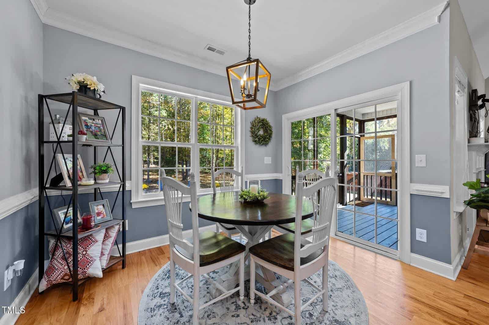 A dining area with a round table and four chairs sits by large windows, offering views of trees. There’s a shelving unit with decor on the left and a sliding glass door leading to a screened porch on the right.