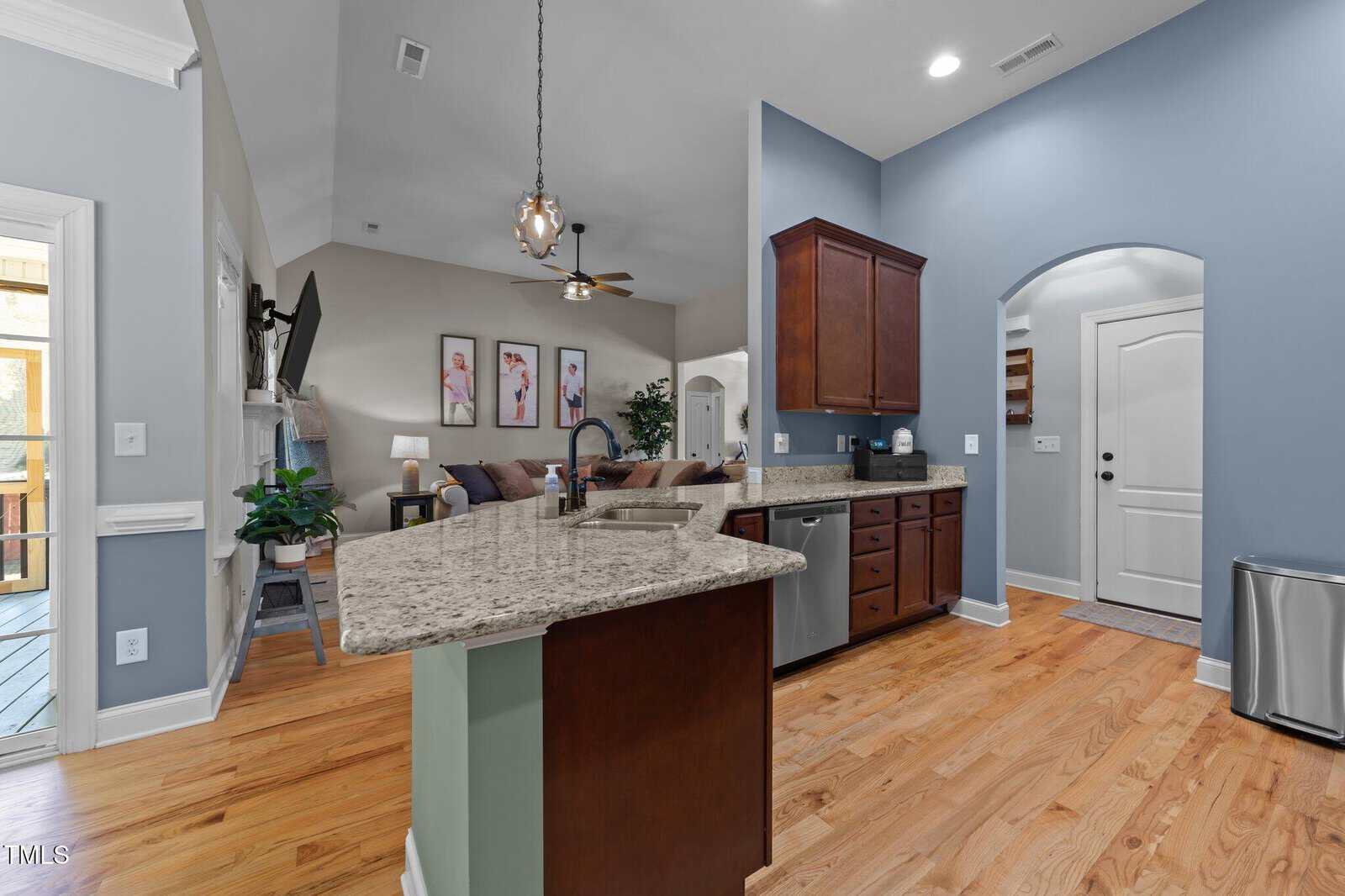 Modern kitchen with granite countertops, stainless steel appliances, wooden cabinets, and hardwood floors. The space opens to a living room with a sofa, TV, ceiling fan, and artwork on the wall.