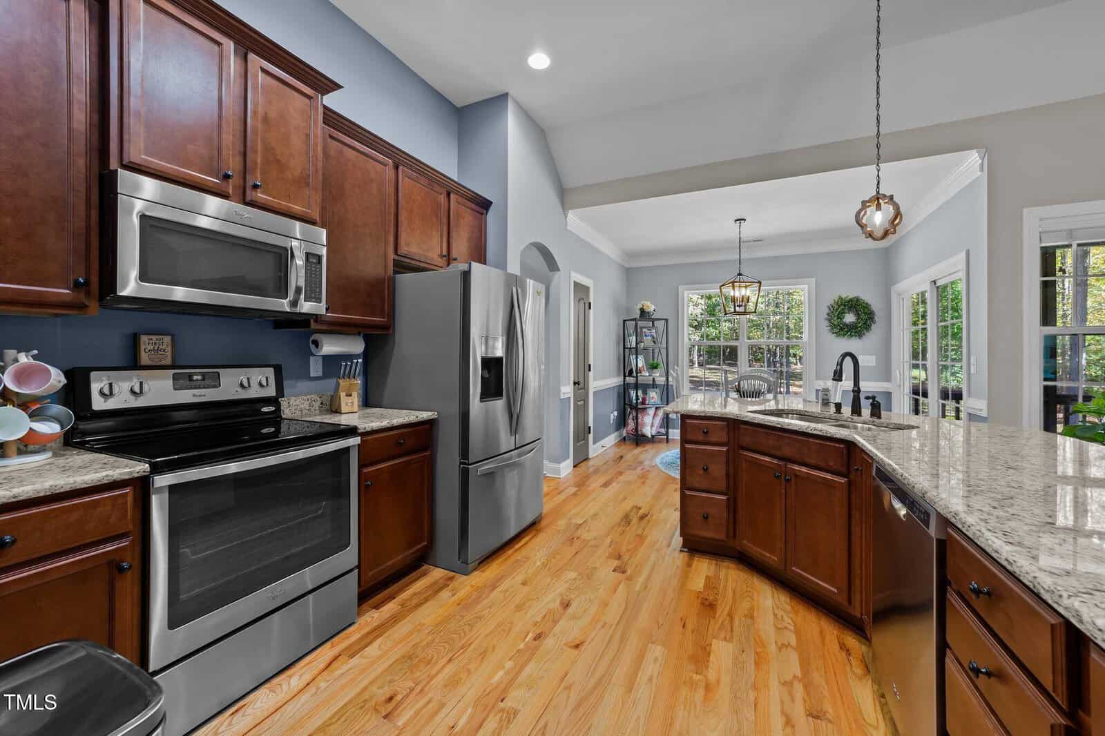Modern kitchen with stainless steel appliances, wooden cabinets, a center island, granite countertops, and hardwood floors. Open layout leads to a bright dining area with large windows and glass doors to the outside.