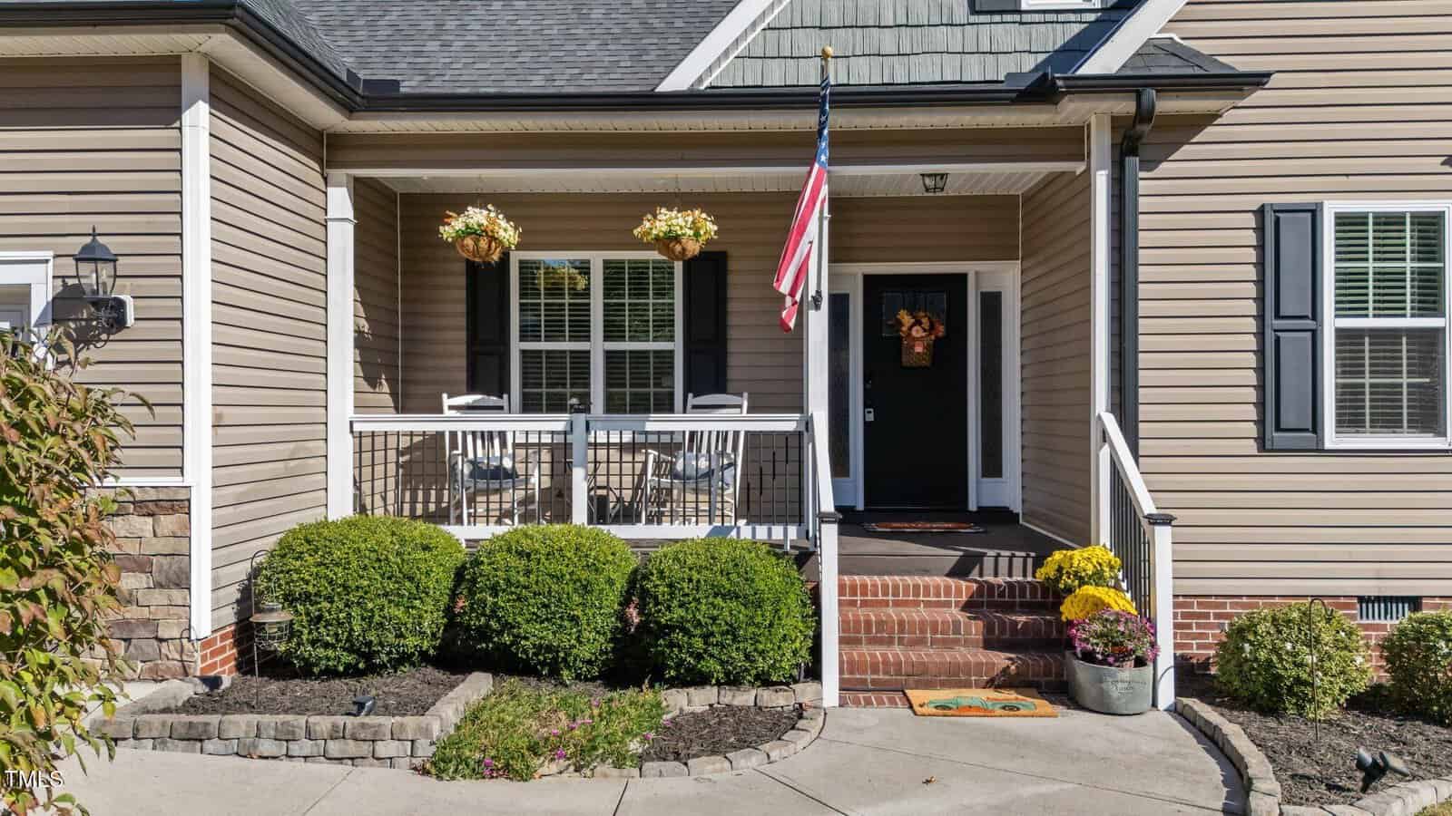 Accessible front porch of a tan house with white trim, two rocking chairs, hanging flower baskets, an American flag, and potted yellow flowers near the steps. Well-maintained bushes border the walkway.