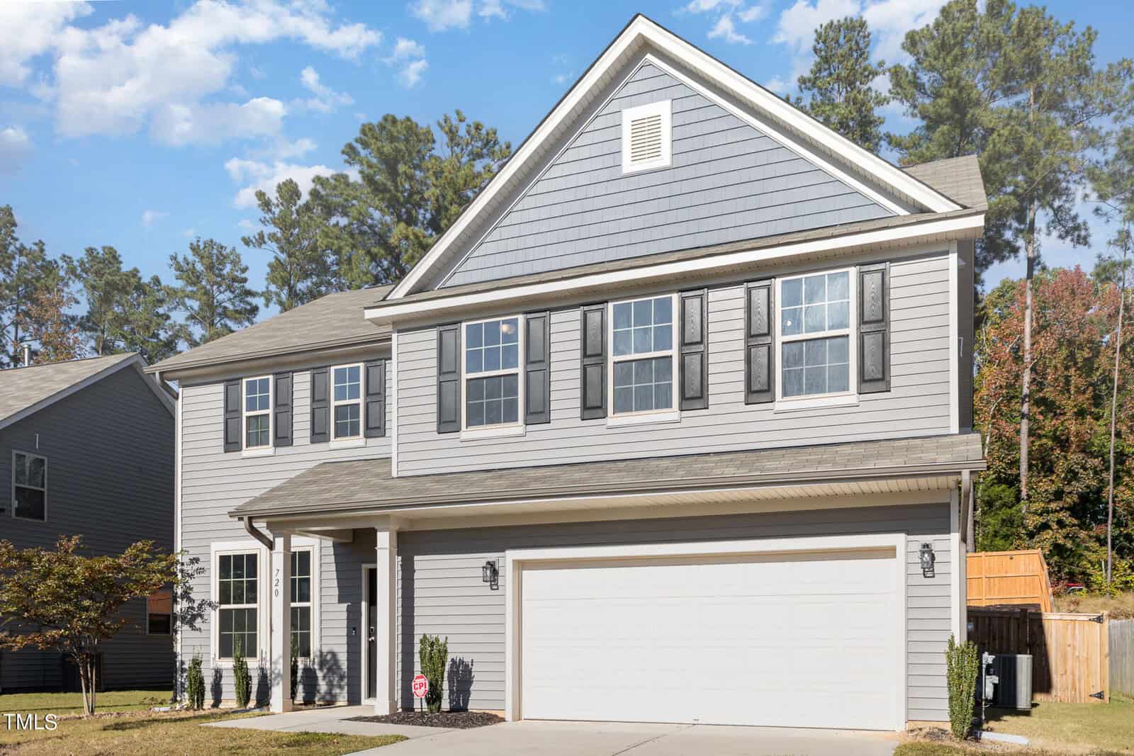 Two-story suburban house with gray siding, black shutters, and a white double garage door. Concrete driveway and small front lawn with a few shrubs. Trees and a wooden fence are visible in the background.