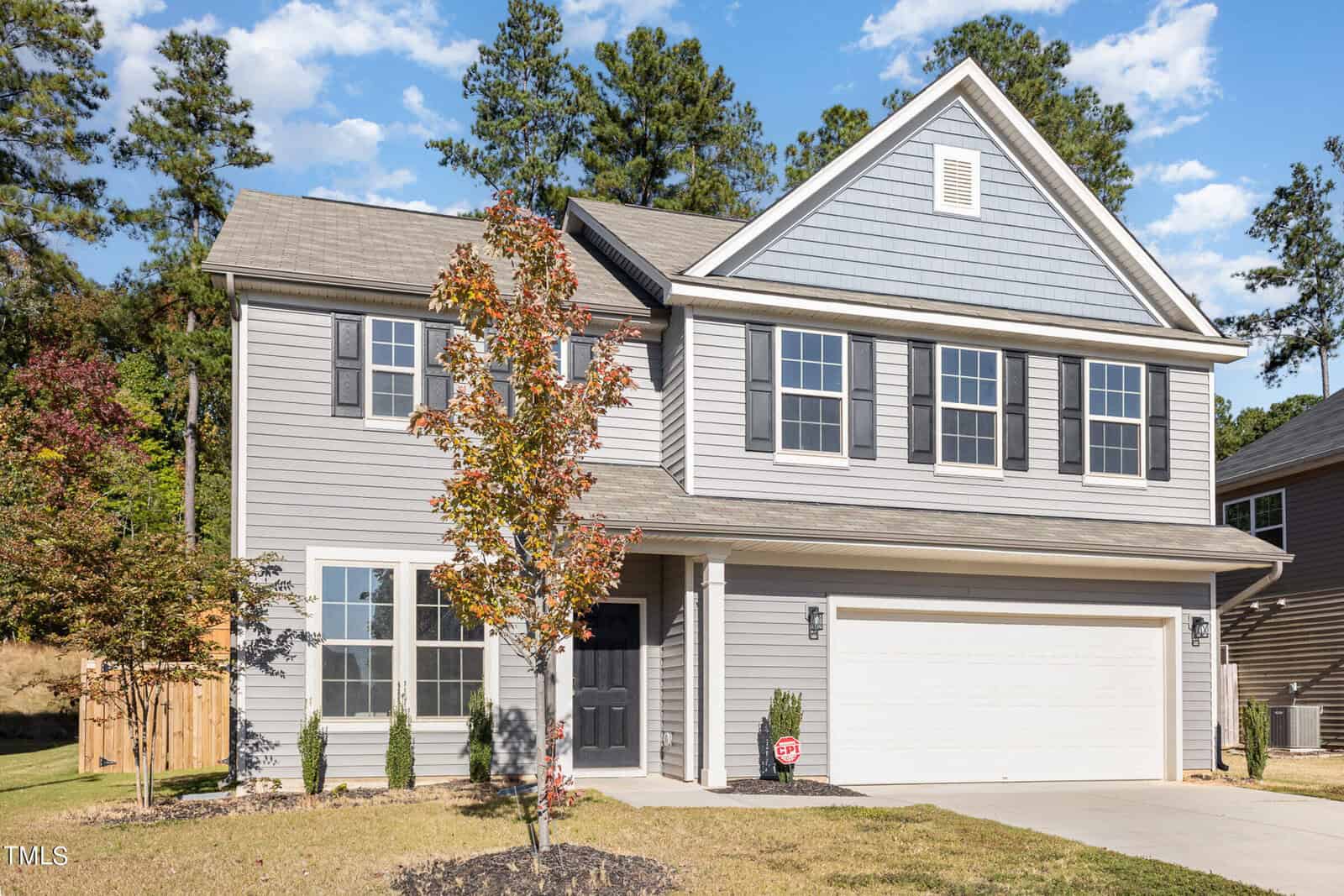 Two-story gray house with black shutters, a covered entryway, and a white double garage. Young trees and shrubs are planted in the front yard. Tall pine trees and blue sky are visible in the background.
