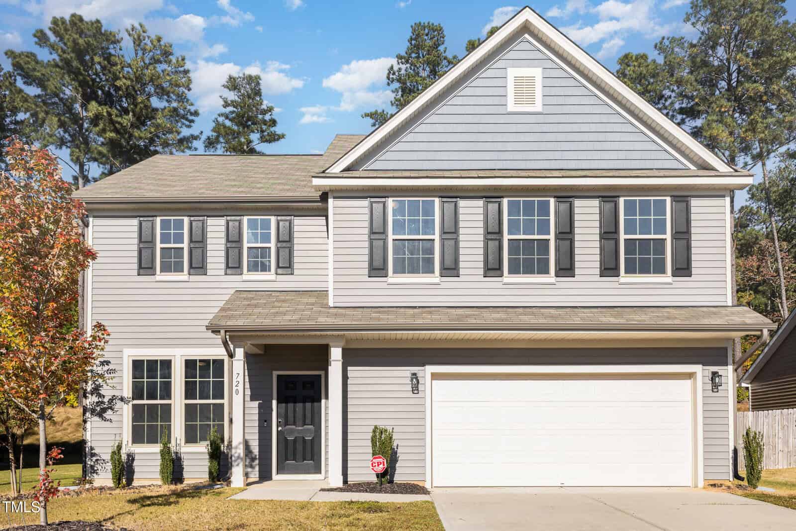 Two-story gray house with white trim, black shutters, and a double garage. There are small shrubs along the front, a concrete driveway, and a young tree on the left side. Tall pine trees are visible in the background.