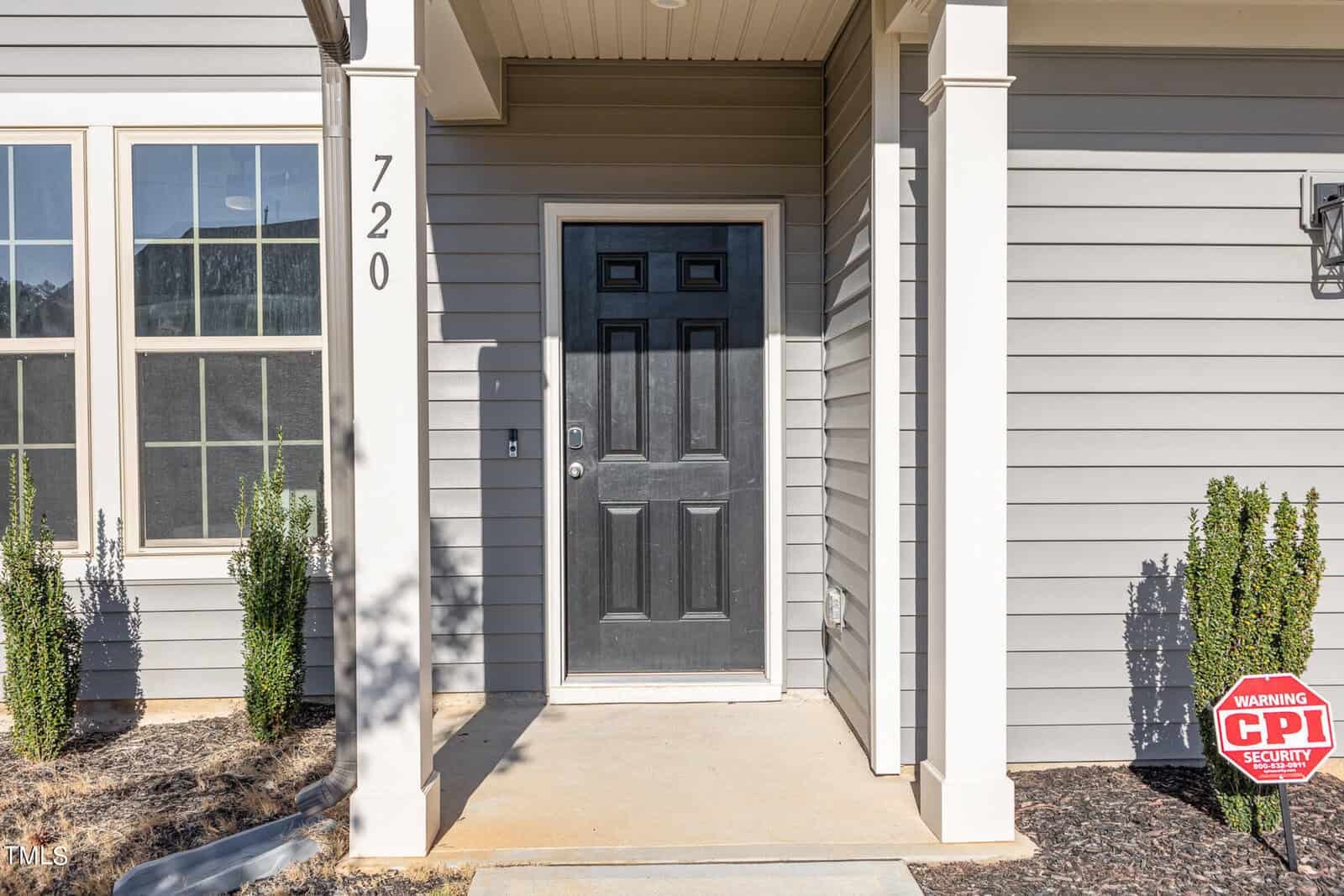 A gray house with white trim, featuring a black front door, the number 720 displayed vertically, small shrubs along the walkway, and a red CPI Security sign in the mulch near the entrance.
