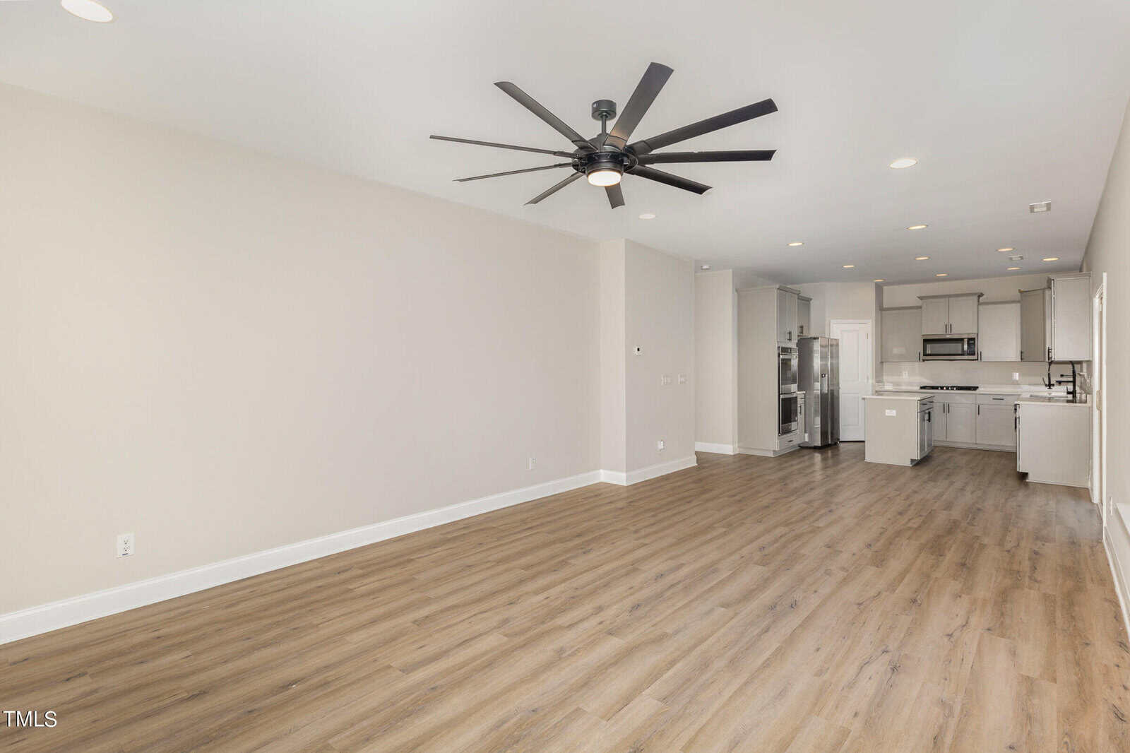 Spacious modern living area with light wood flooring, white walls, large black ceiling fan, and recessed lighting, leading to an open kitchen with stainless steel appliances and white cabinetry.