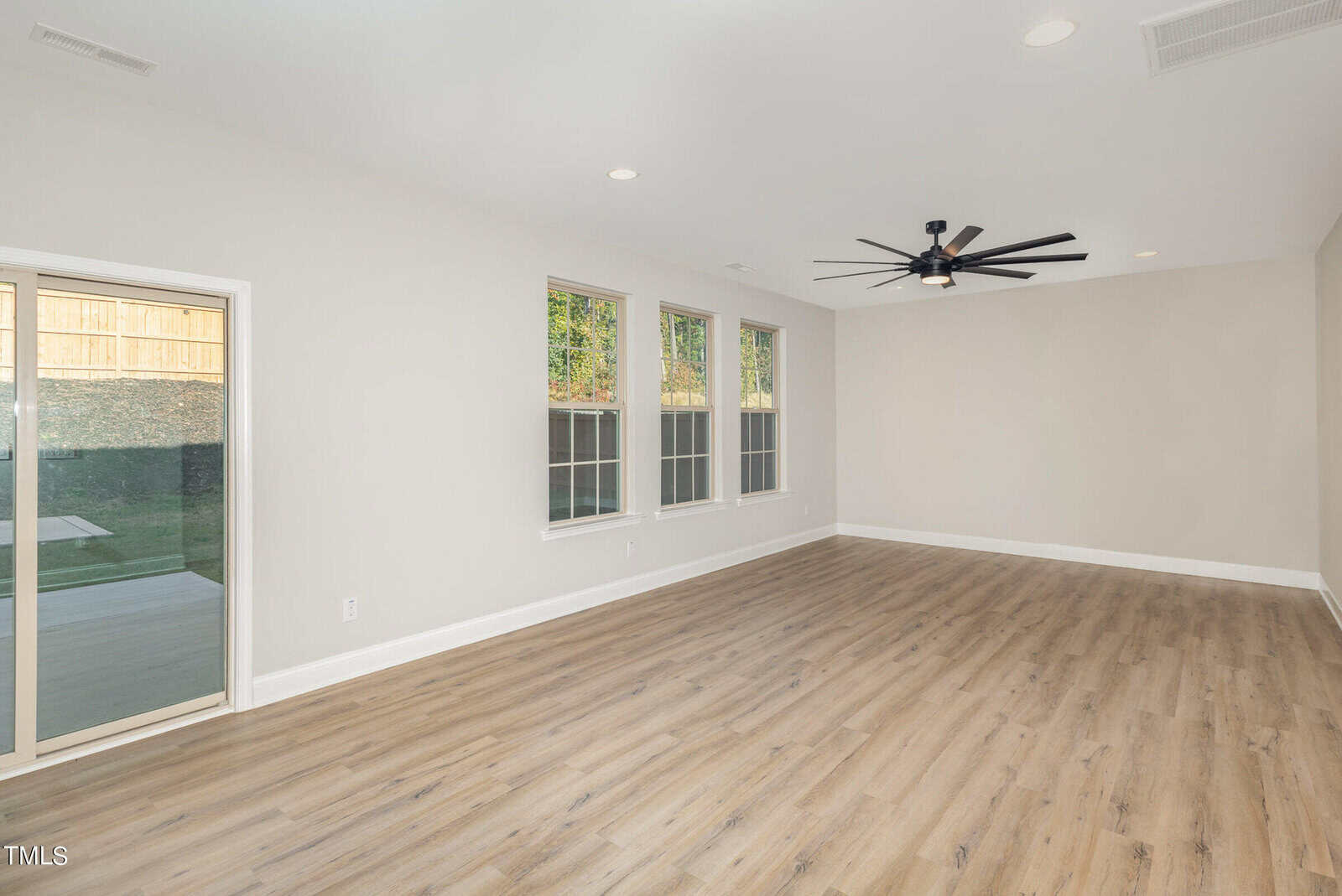 A spacious, empty room with light wood flooring, beige walls, three large windows, a black ceiling fan, recessed lighting, and a sliding glass door leading to a backyard.