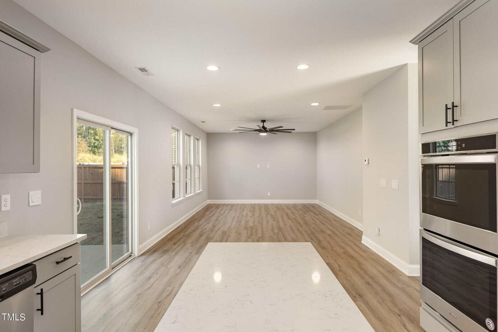Open-concept kitchen and living area with light wood flooring, white walls, recessed lighting, large windows, sliding glass door, a ceiling fan, built-in double ovens, and a kitchen island with a white countertop.