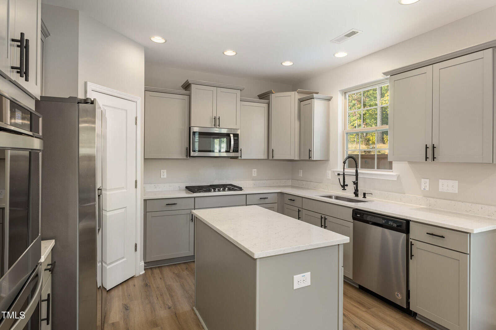 Modern kitchen with light gray cabinets, stainless steel appliances, a center island, and wood flooring. There is a window above the sink letting in natural light and recessed lighting in the ceiling.