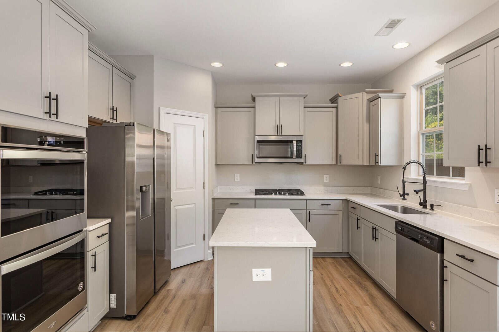 Modern kitchen with light gray cabinets, stainless steel appliances including double ovens, a refrigerator, and dishwasher, a central island with a white countertop, and wood-style flooring. Window above the sink lets in natural light.