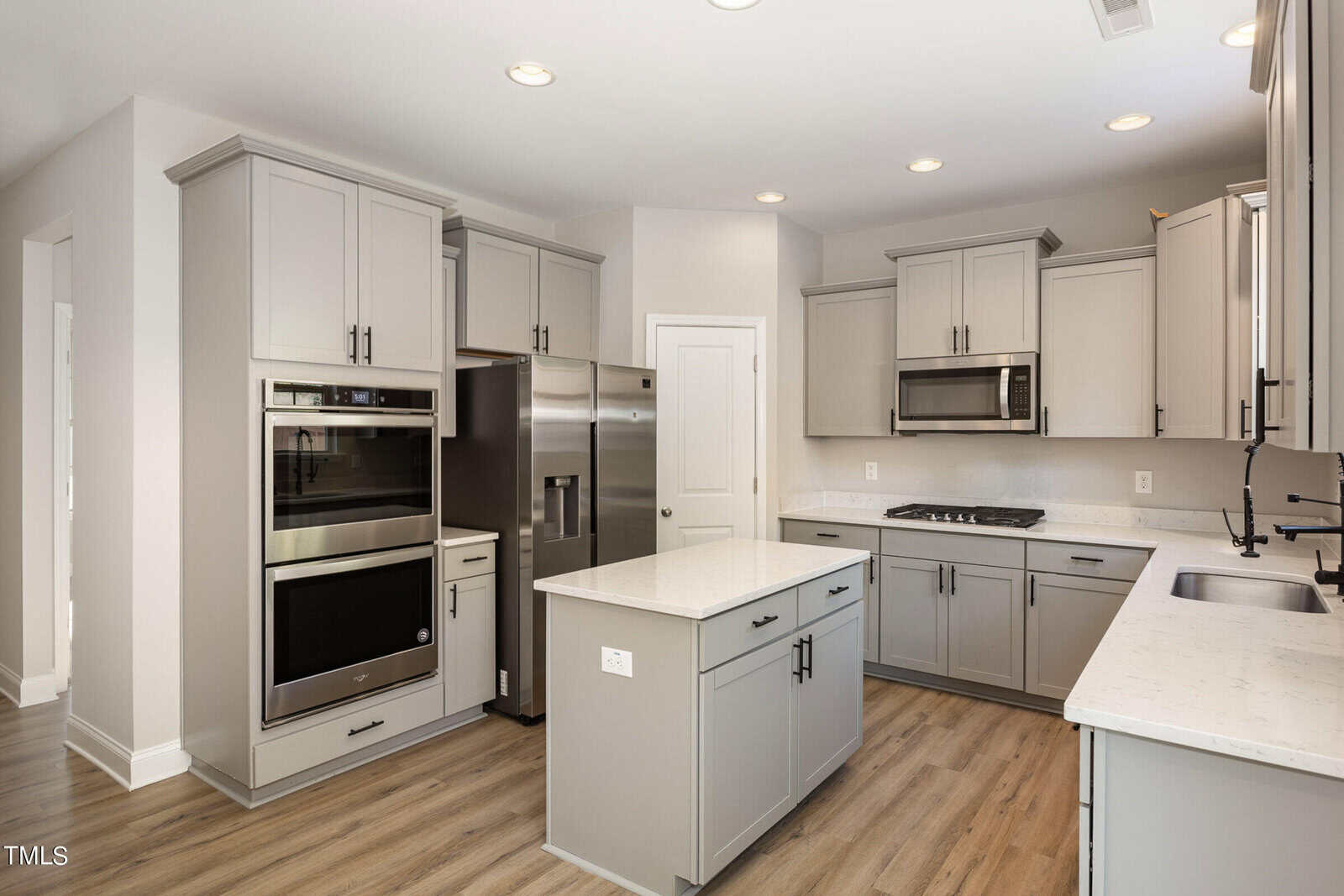 Modern kitchen with light gray cabinets, stainless steel appliances, a small central island, white countertops, wood-style flooring, recessed ceiling lights, and a double wall oven.