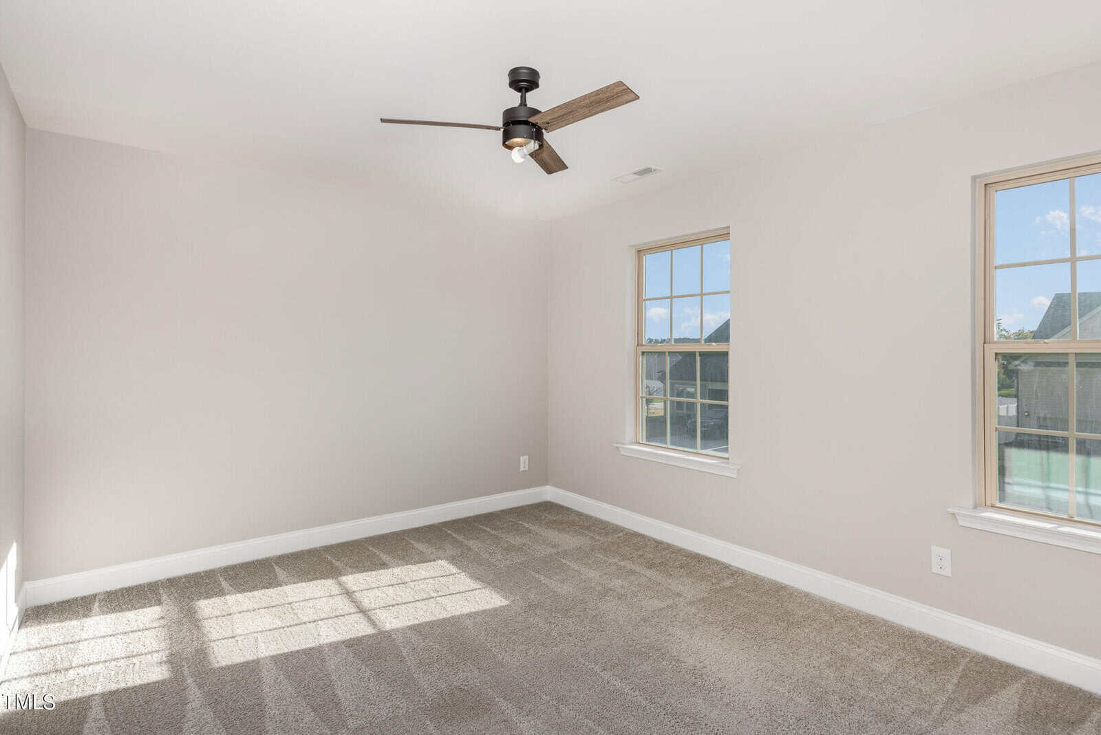 Bright, empty room with beige carpet, light gray walls, two windows, a black ceiling fan, and white trim. Sunlight streams in through the windows, creating shadows on the carpet.