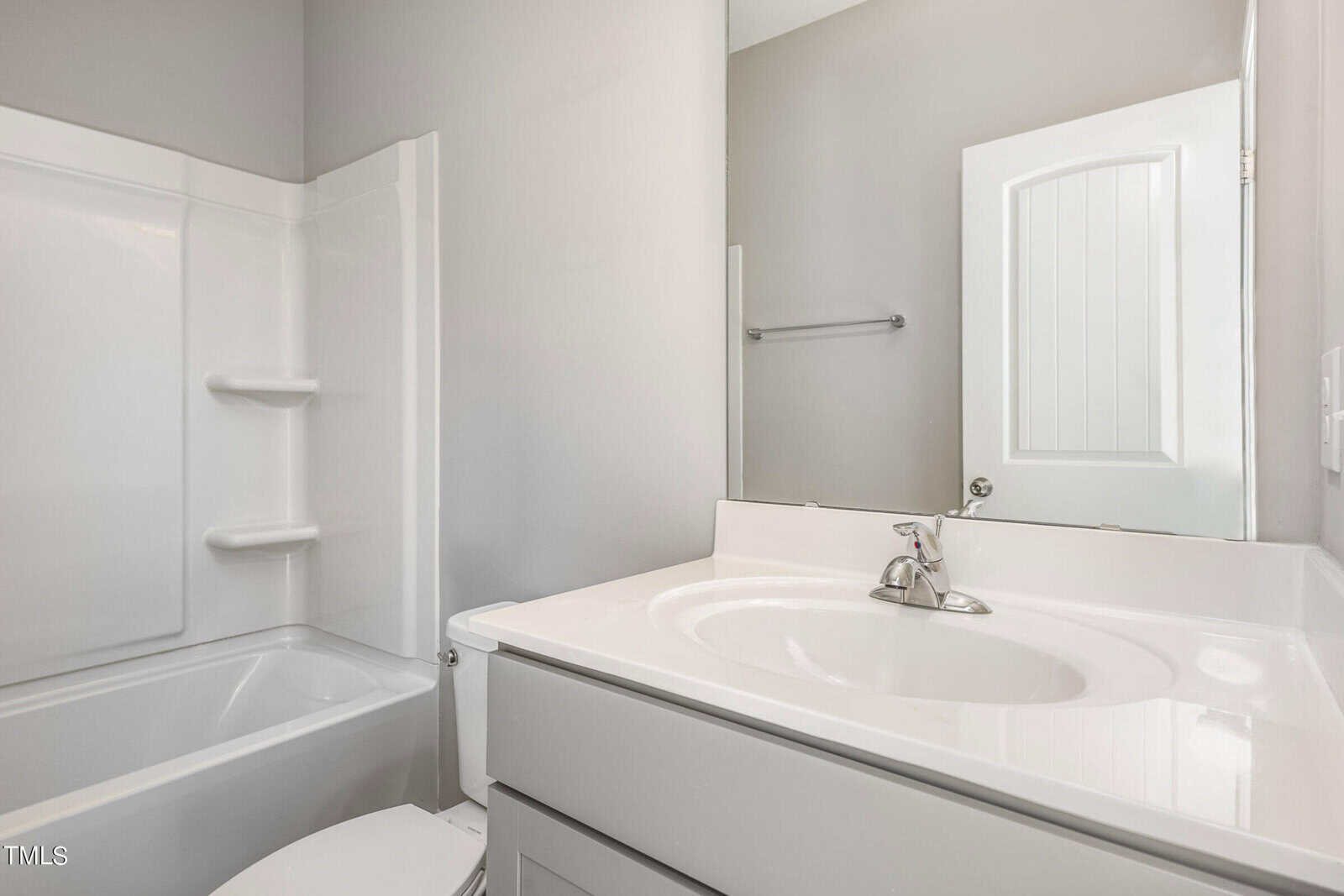 Bright bathroom with a white bathtub-shower combo, white toilet, light gray vanity with sink, chrome faucet, large mirror, and a white paneled door. Walls are painted a light gray shade.