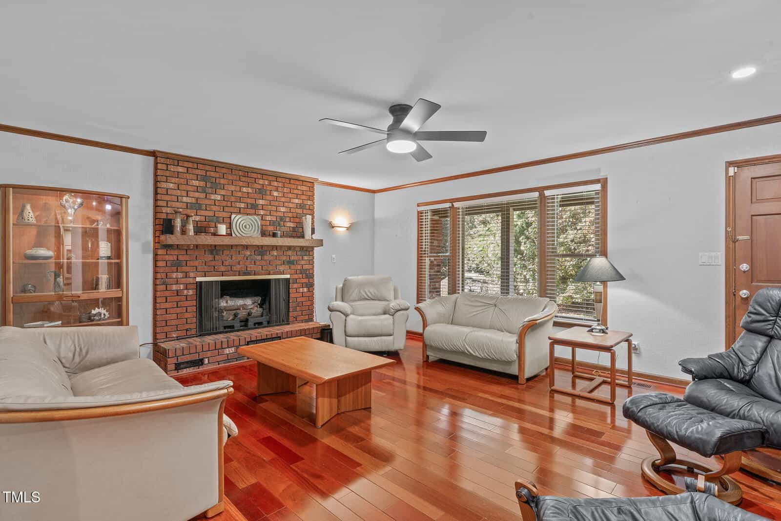Living room with wood floors, brick fireplace, light-colored sofas, a wooden coffee table, display case, recliner, large window with blinds, and a ceiling fan with lights.