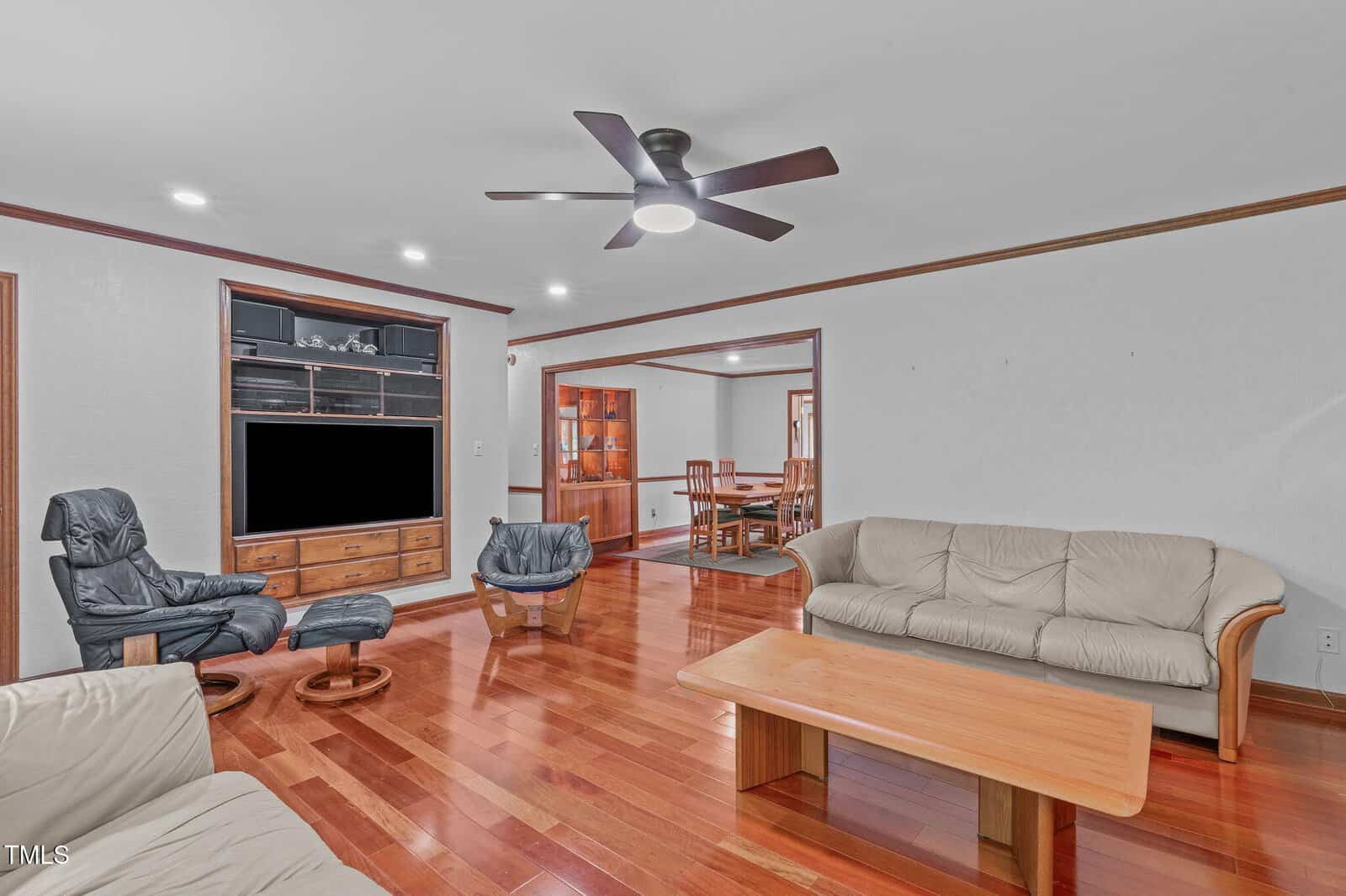 A living room with wood floors, a beige leather sofa set, a light wood coffee table, a reclining chair with ottoman, a TV in a wooden entertainment center, and a ceiling fan. Dining area visible through an open doorway.