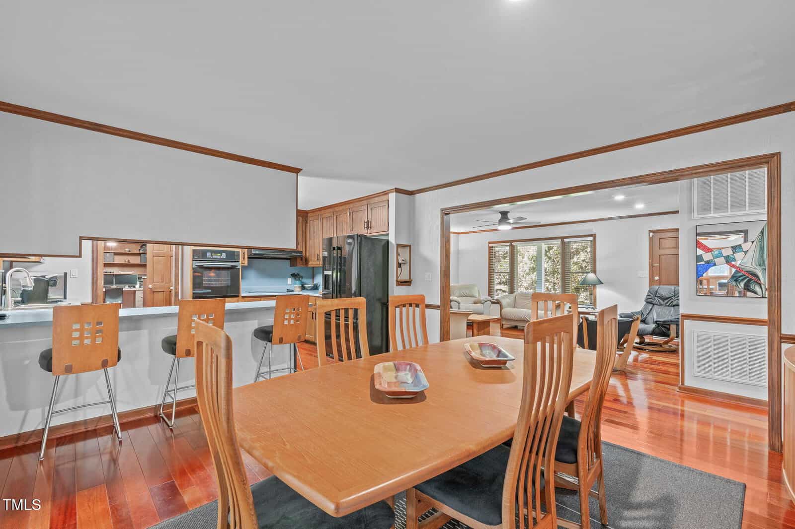 Open-concept dining area with a wooden table and chairs, adjacent to a kitchen with barstools and an extended countertop. The space leads to a living room with armchairs and large windows.