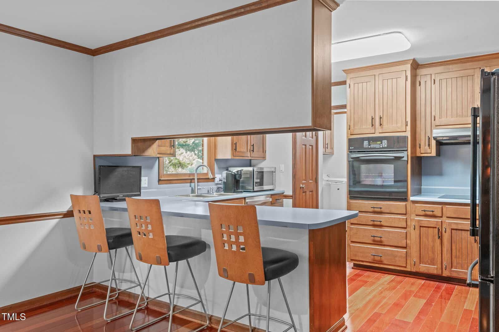 Modern kitchen with wood cabinets, a breakfast bar with three chairs, built-in oven, black refrigerator, microwave, and a laptop on the counter. The space features red-toned hardwood floors and white countertops.