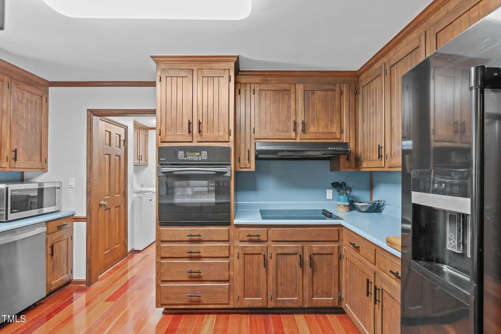 Kitchen with wooden cabinets, black appliances, blue countertops, and red-toned wood floors. A potted plant and cutting board are on the counter. A laundry room is visible through an open doorway.
